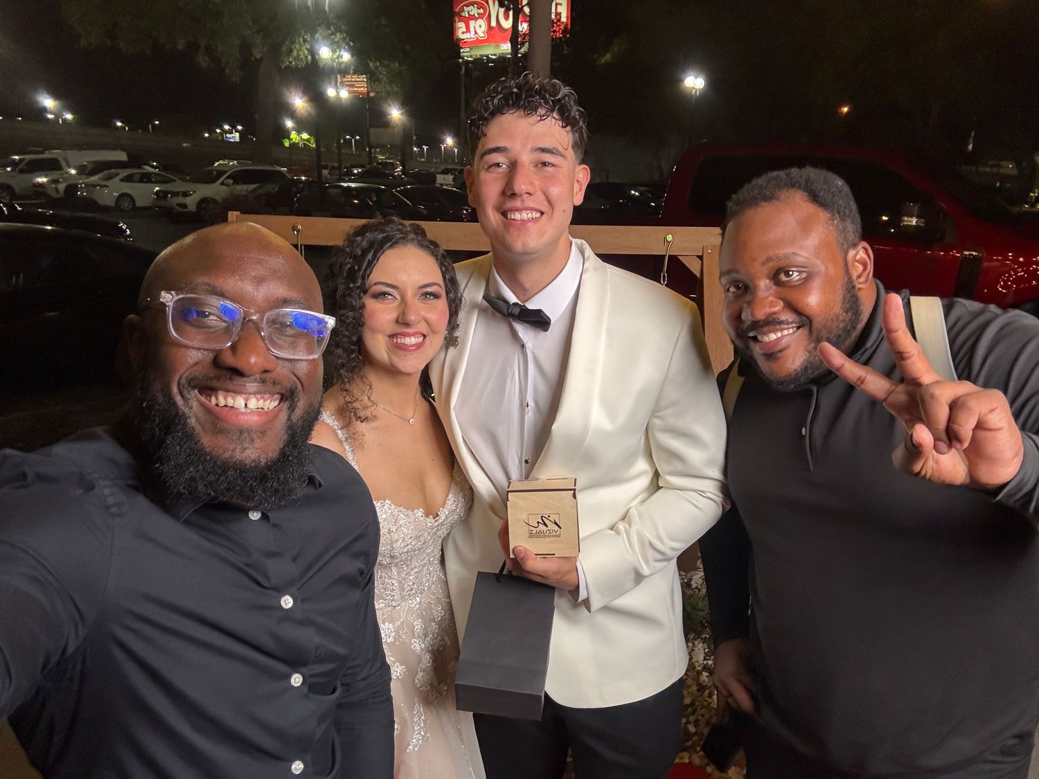 Four friends pose together outdoors at night with one holding an award box while celebrating an occasion.
