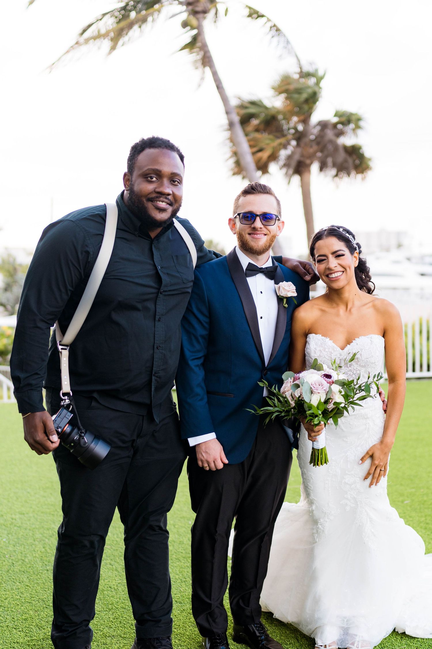 Wedding photographer in black jacket poses with newlyweds on a grassy lawn with palm trees swaying in the background.