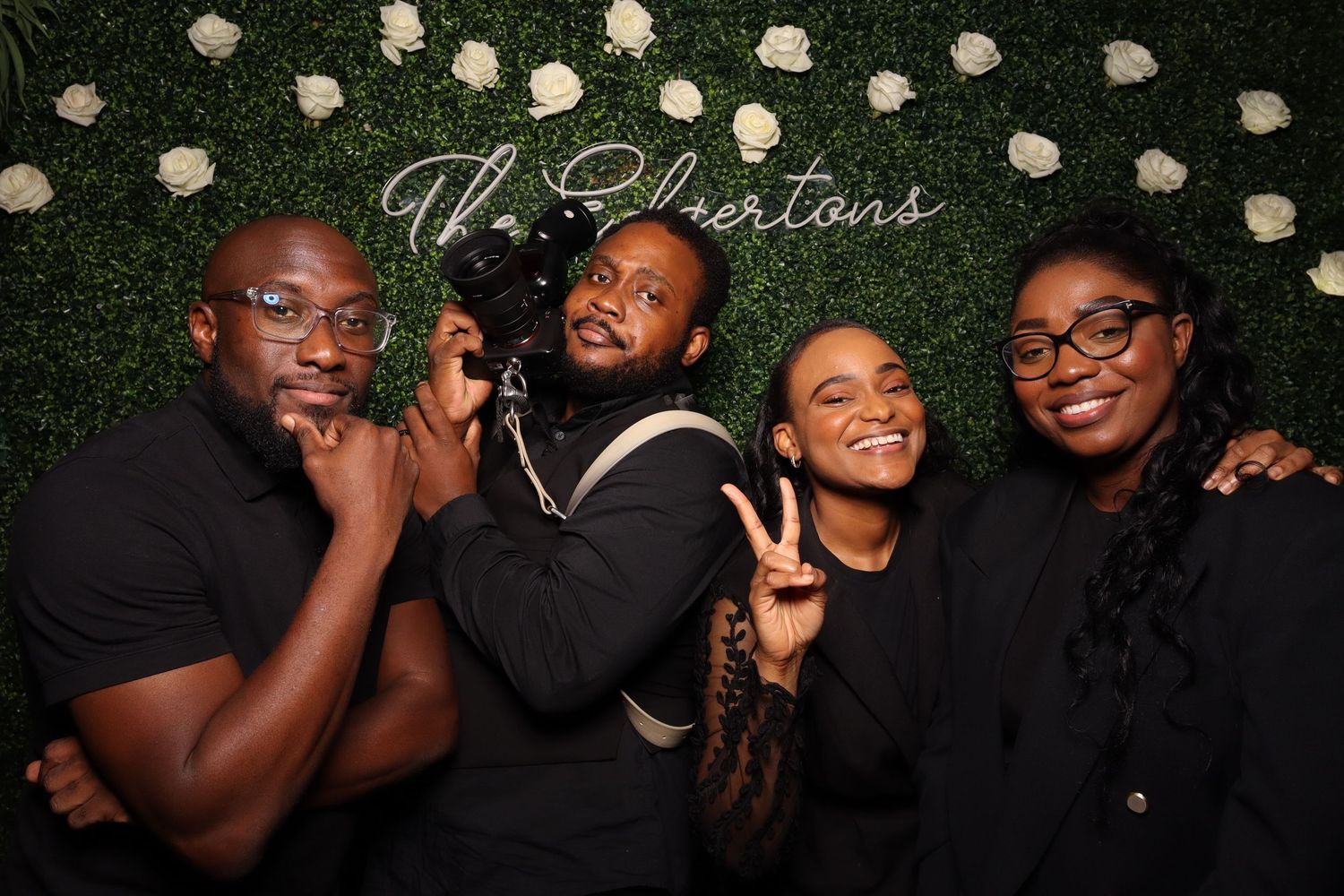 Group photo in front of greenery wall with white flowers, showing four people in black outfits posing playfully together.