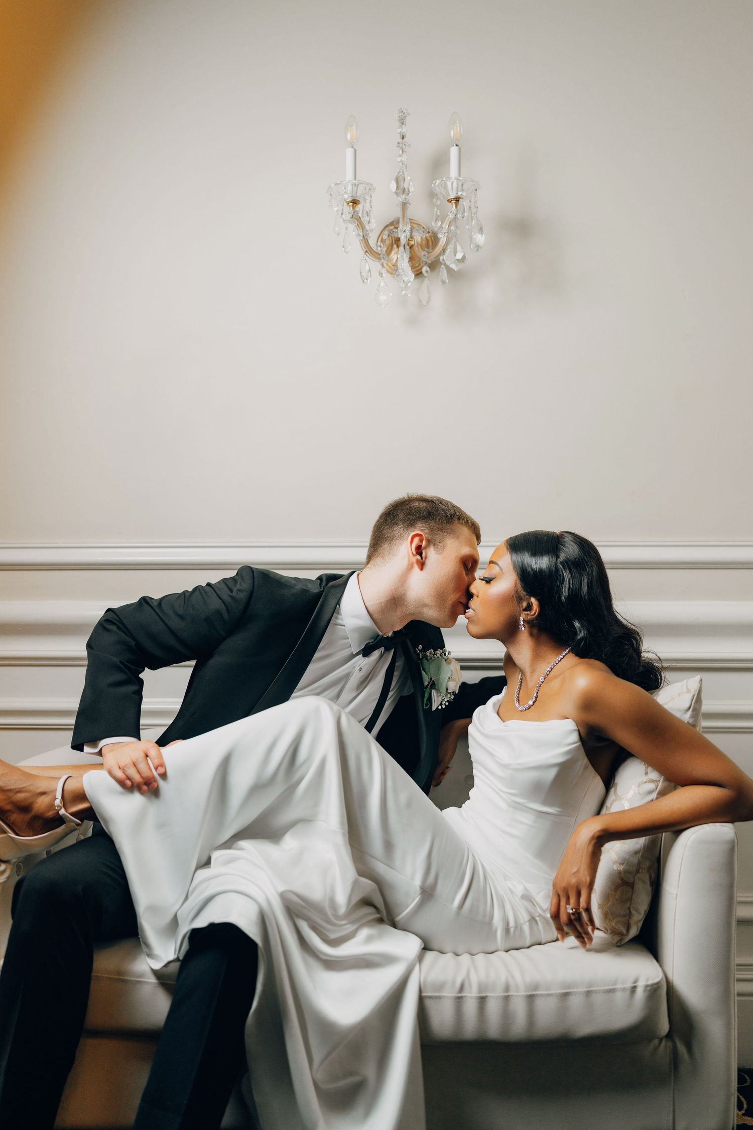 A romantic wedding couple shares an intimate moment on an elegant white sofa beneath a crystal chandelier.