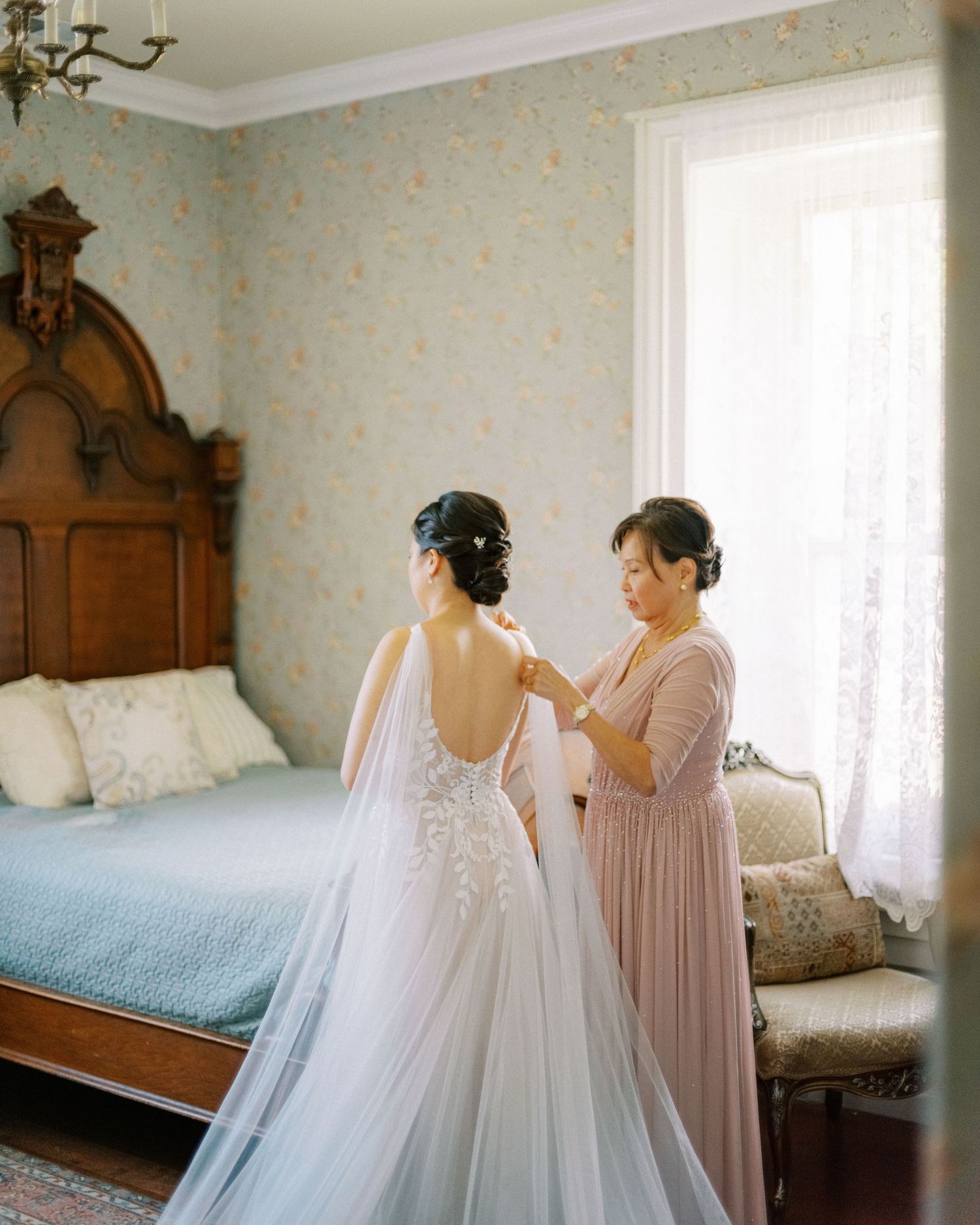 Wedding gown and bridesmaid dress hanging in a sunlit bedroom with antique wooden furniture and floral wallpaper.