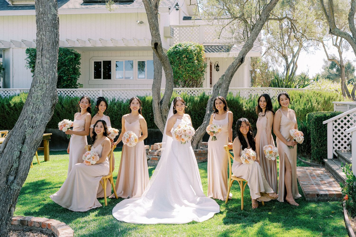 Wedding party in champagne-colored dresses poses outdoors under trees in a garden setting.