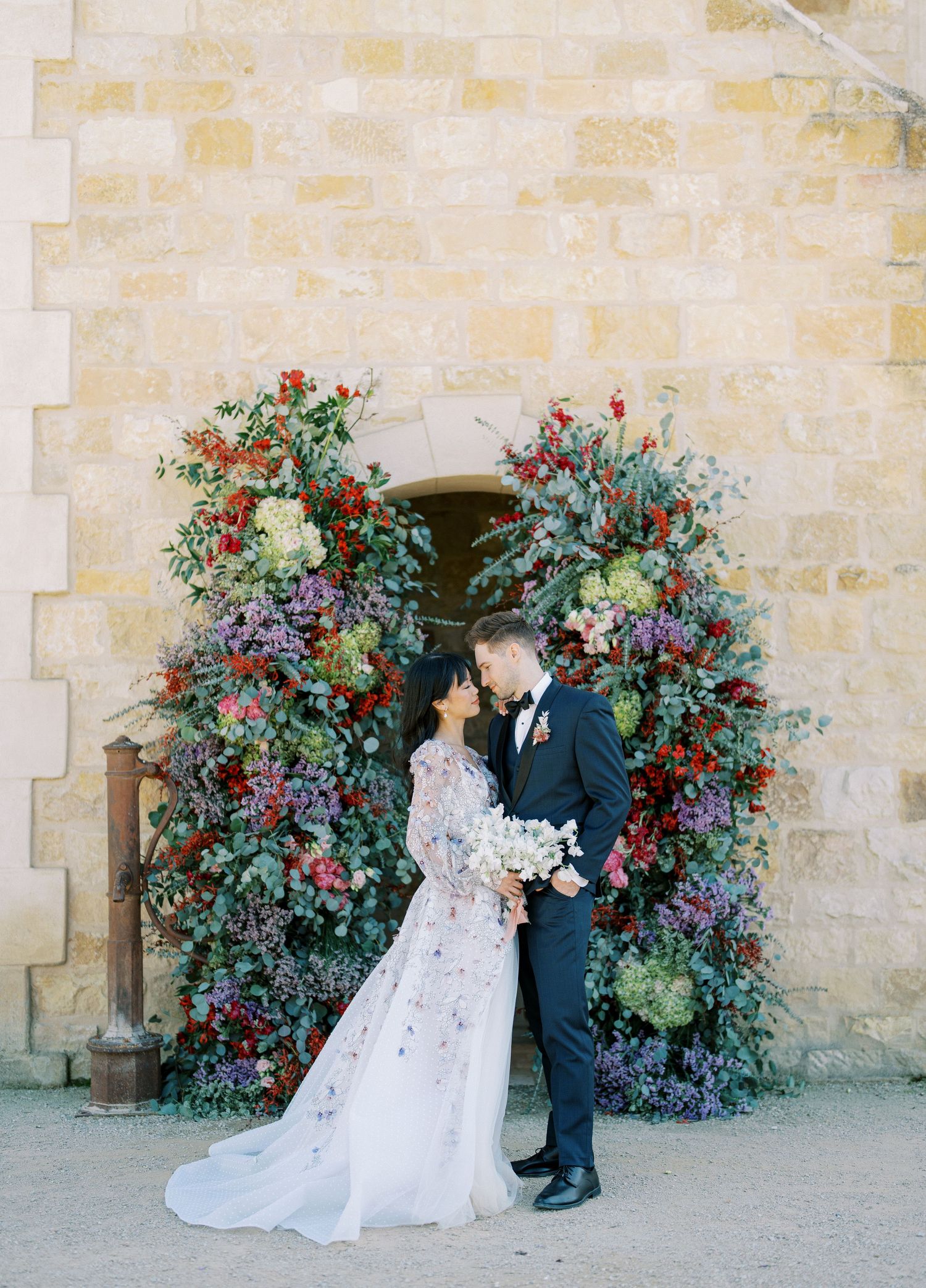 A couple stands in front of an ornate floral arch with blue and red blooms against a stone wall at their wedding.