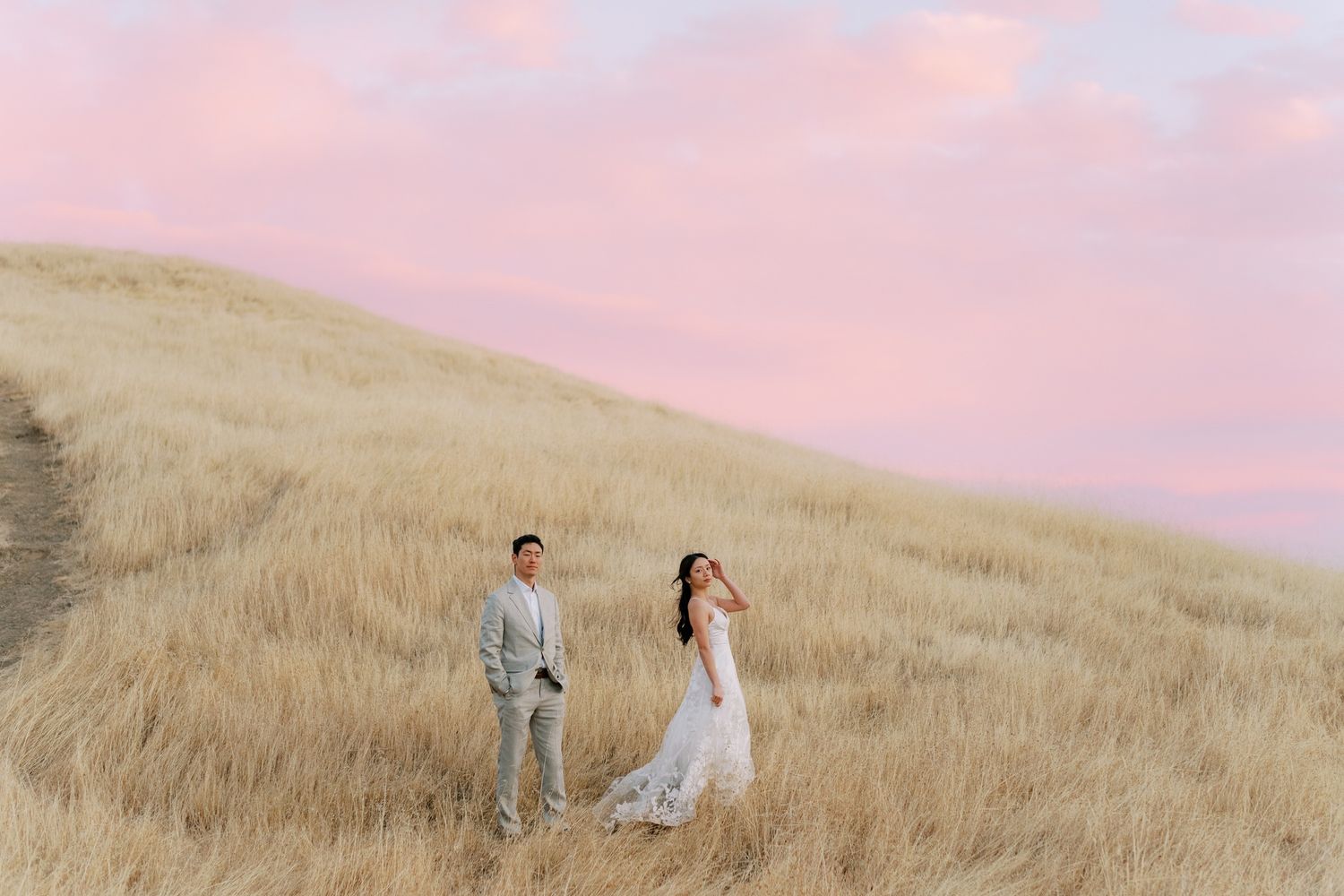 A couple poses together on a golden hillside during sunset while wearing elegant wedding attire.