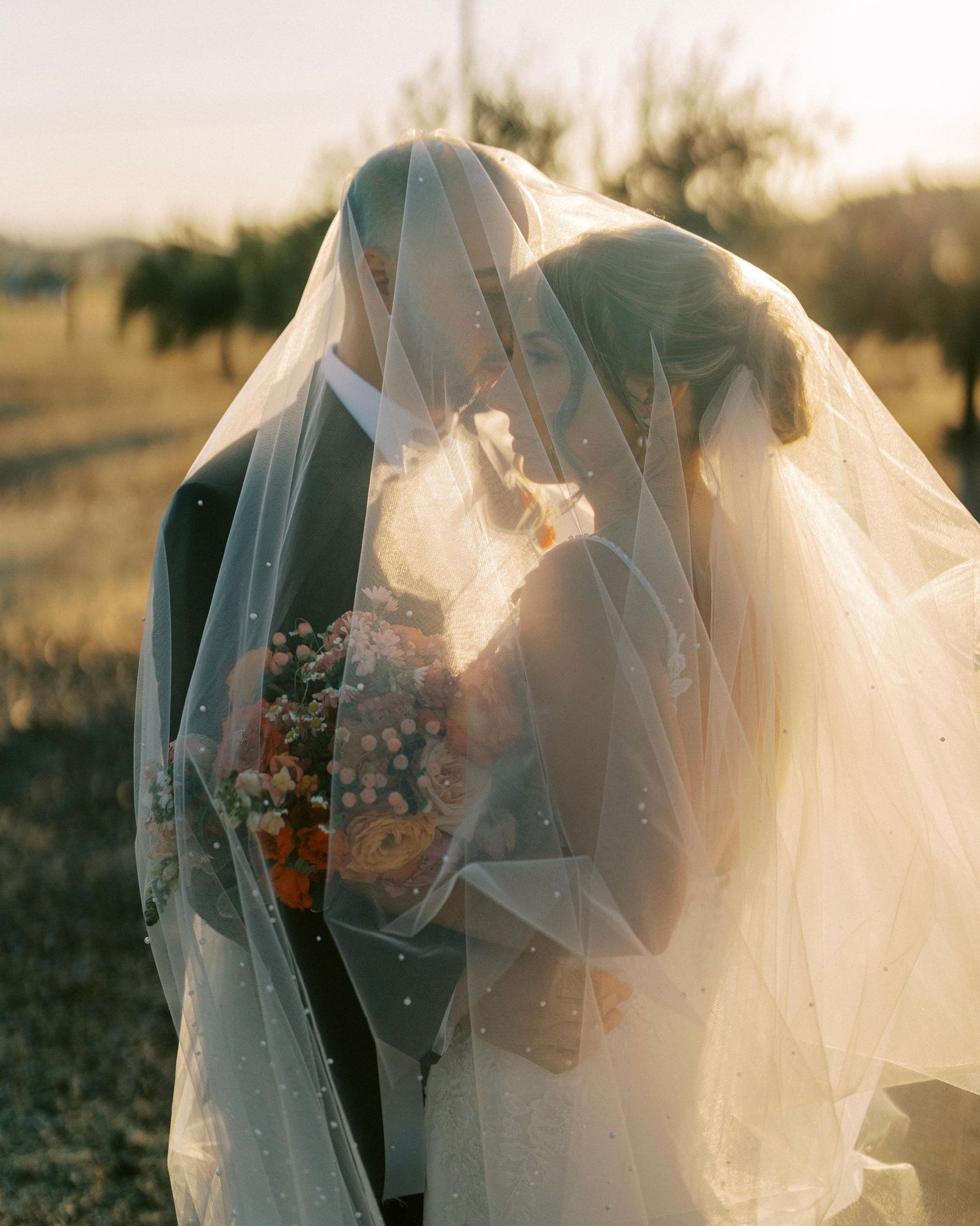 A romantic sunset silhouette of a couple in wedding attire embracing outdoors with a flowing veil in the golden light.