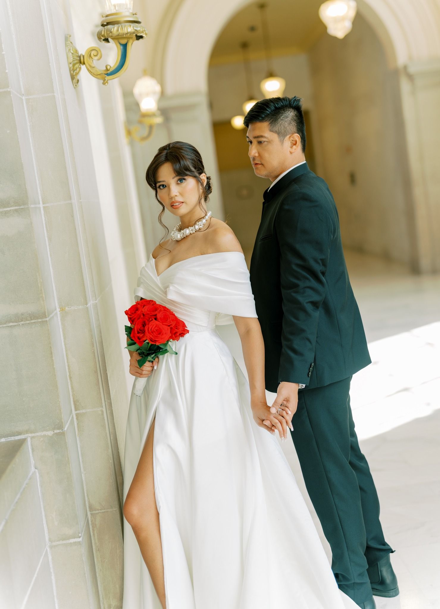 Elegant wedding couple poses in a sunlit hallway with white columns and warm lighting creating romantic shadows.