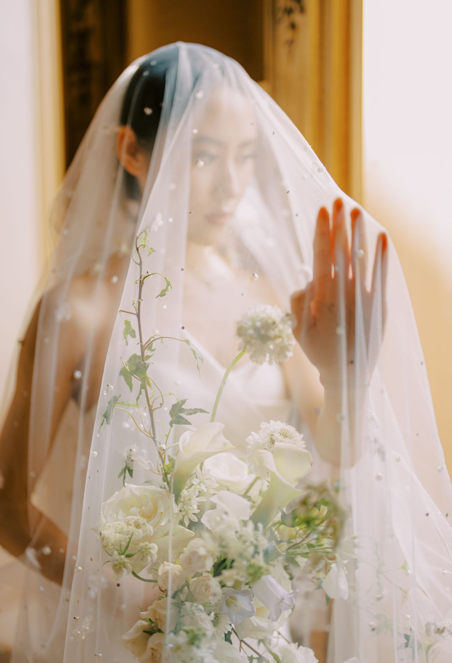 A bride in a white veil holds an elegant bouquet of white flowers and greenery against a soft, warm background.