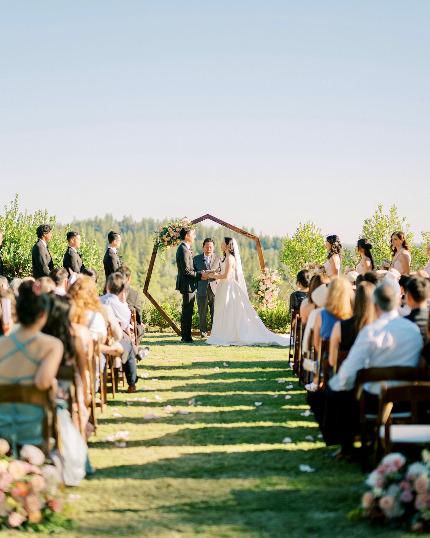 Outdoor wedding ceremony in a scenic field with guests seated on wooden chairs surrounded by greenery.