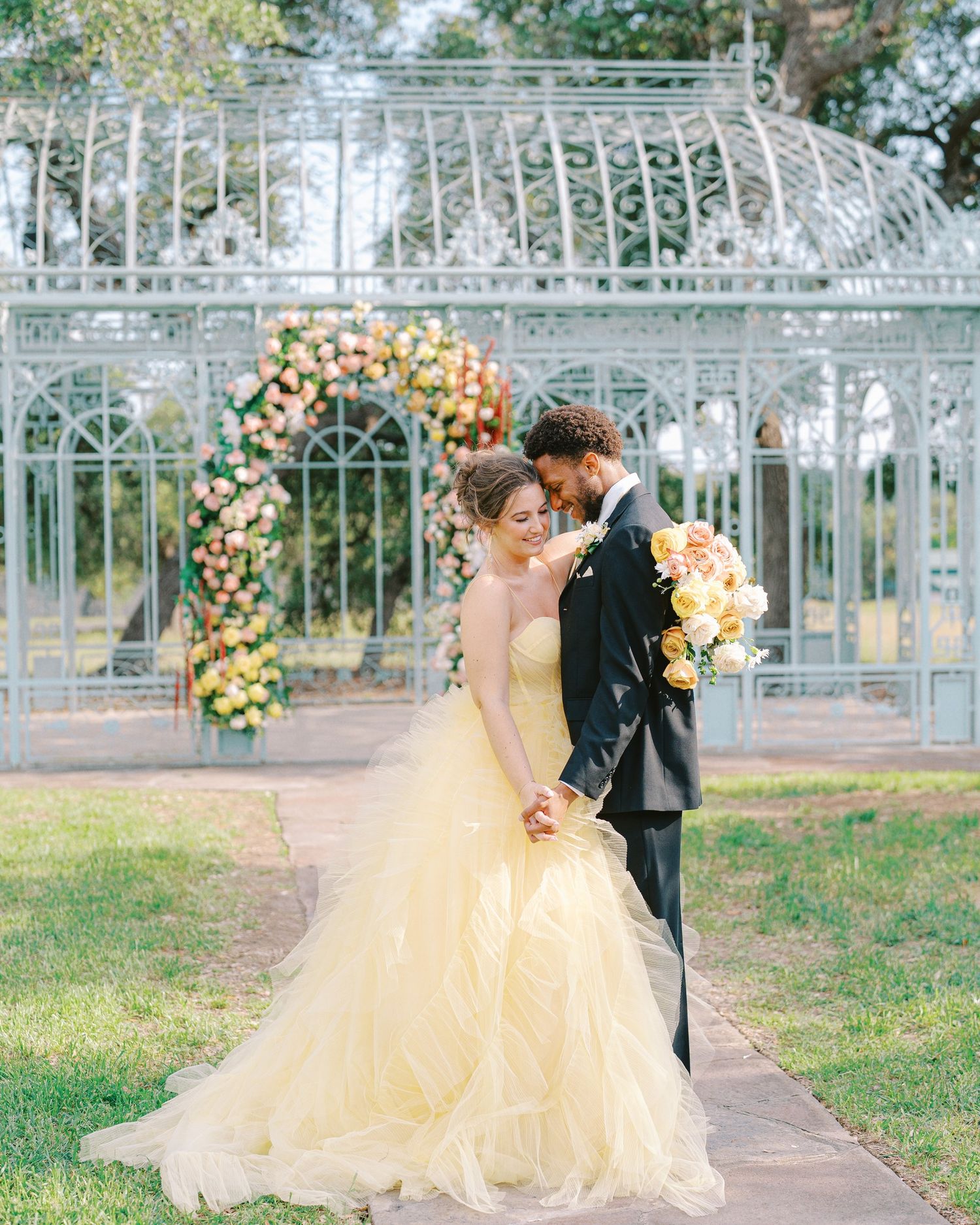 A romantic wedding couple shares intimate moments in a garden setting with white gates and floral decorations.