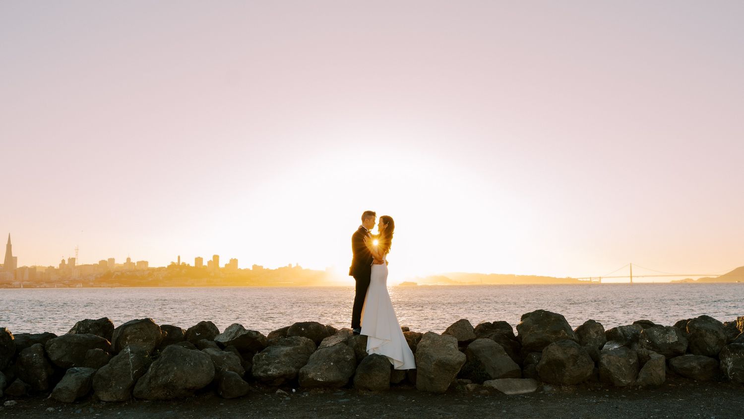 Silhouetted couple embraces on rocky beach against orange sunset sky with city skyline in background.