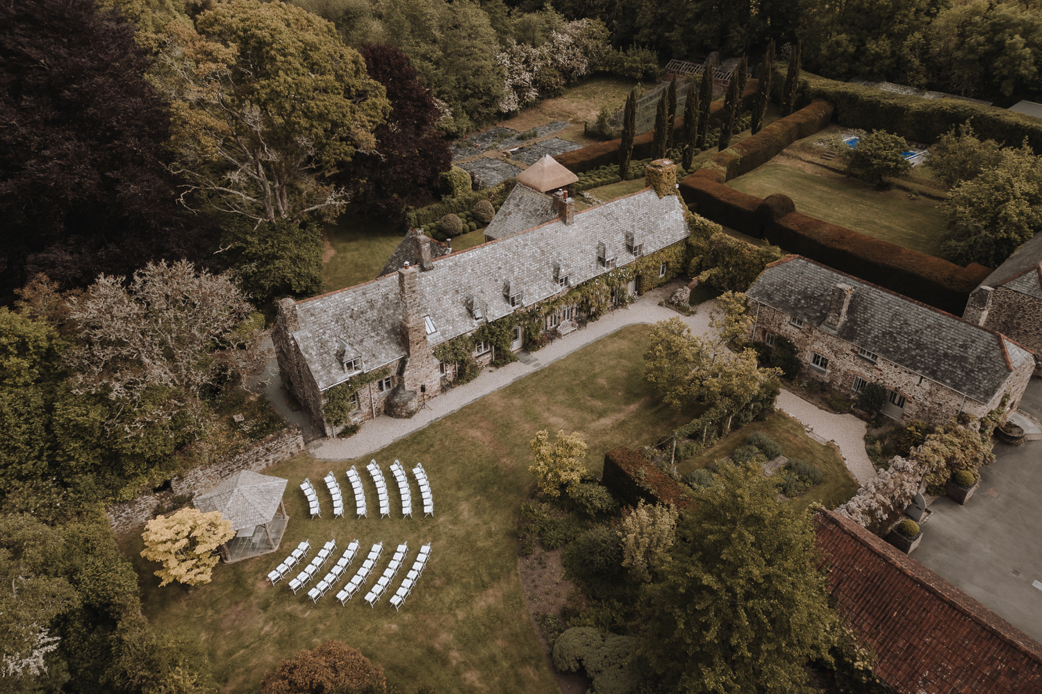 Aerial drone shot of The Great Barn Exeter, ceremony setup with white chairs arranged on manicured lawn surrounded by trees.