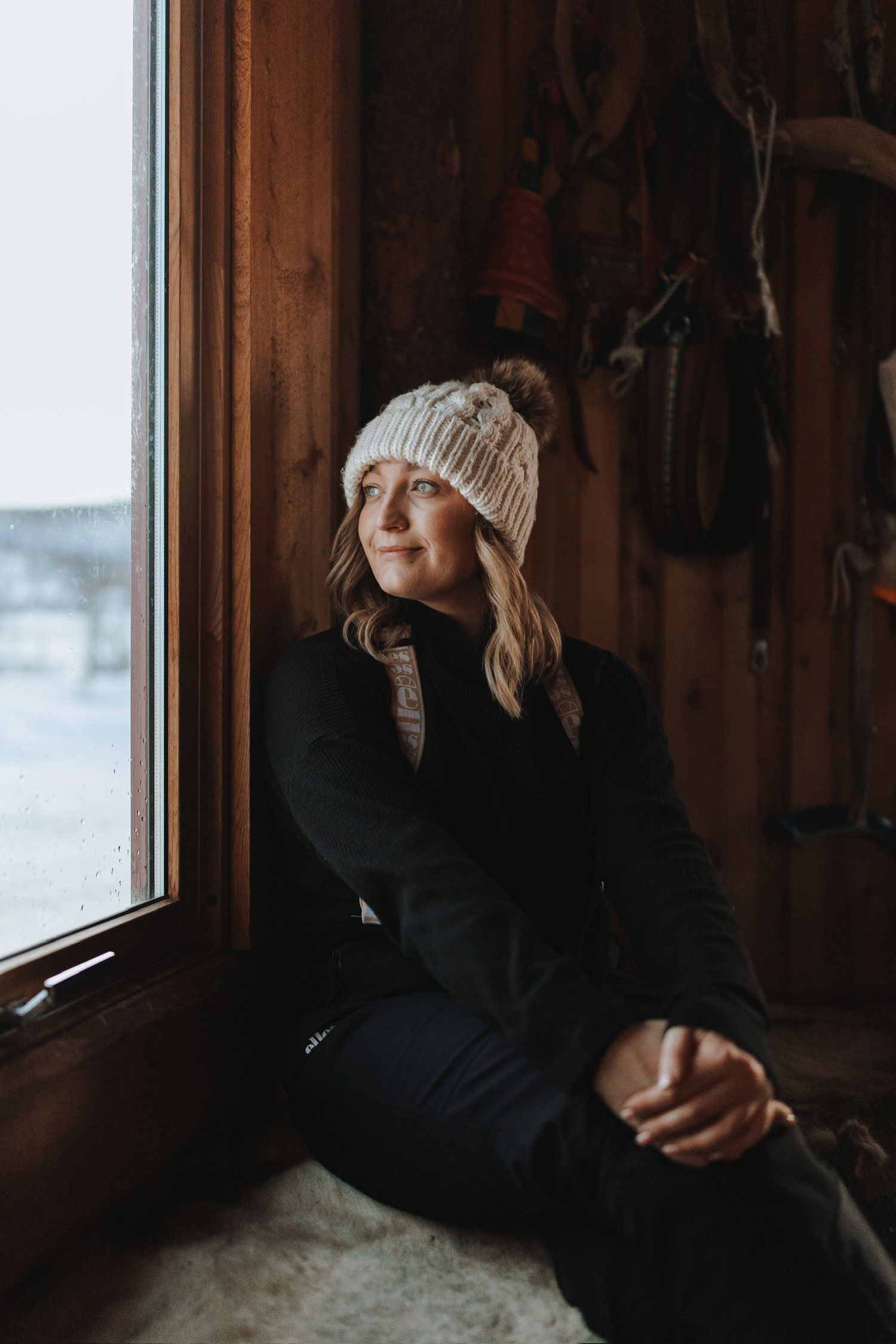 Person in knit beanie and black sweater gazes thoughtfully out cabin window on winter day.