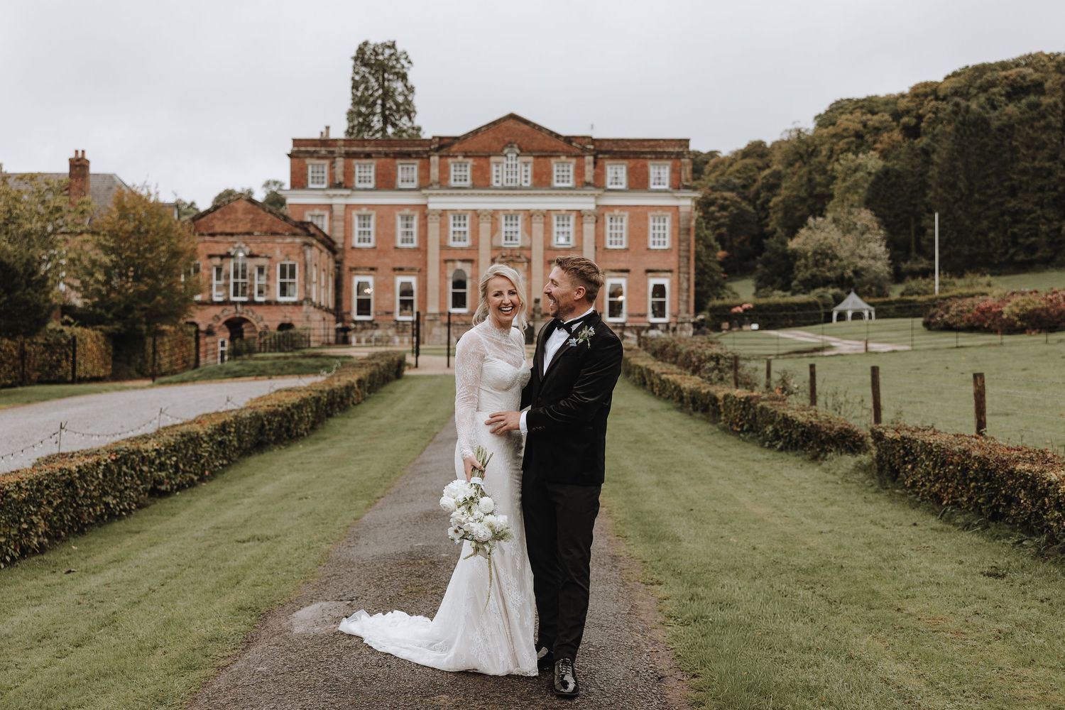 A couple in formal wedding attire walks together on a landscaped path outside a stately brick mansion.