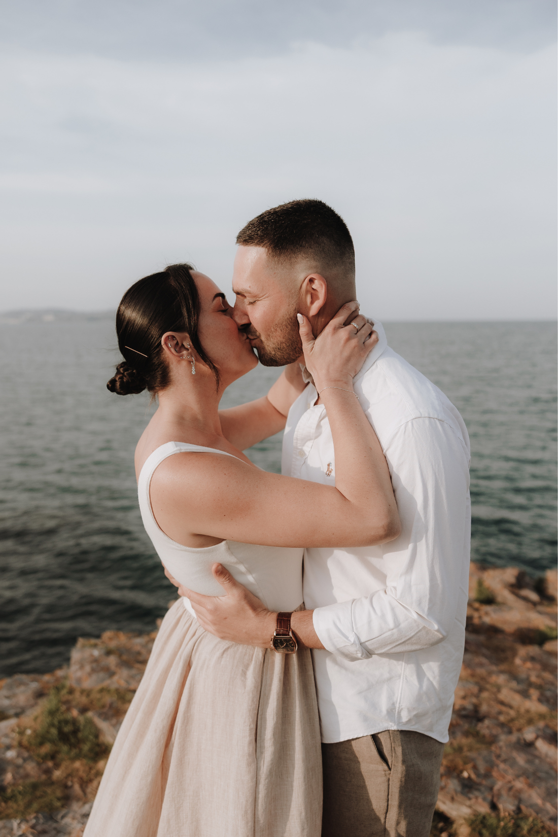 A couple shares a romantic kiss by the ocean, dressed in elegant white and beige outfits on rocky coastal terrain.