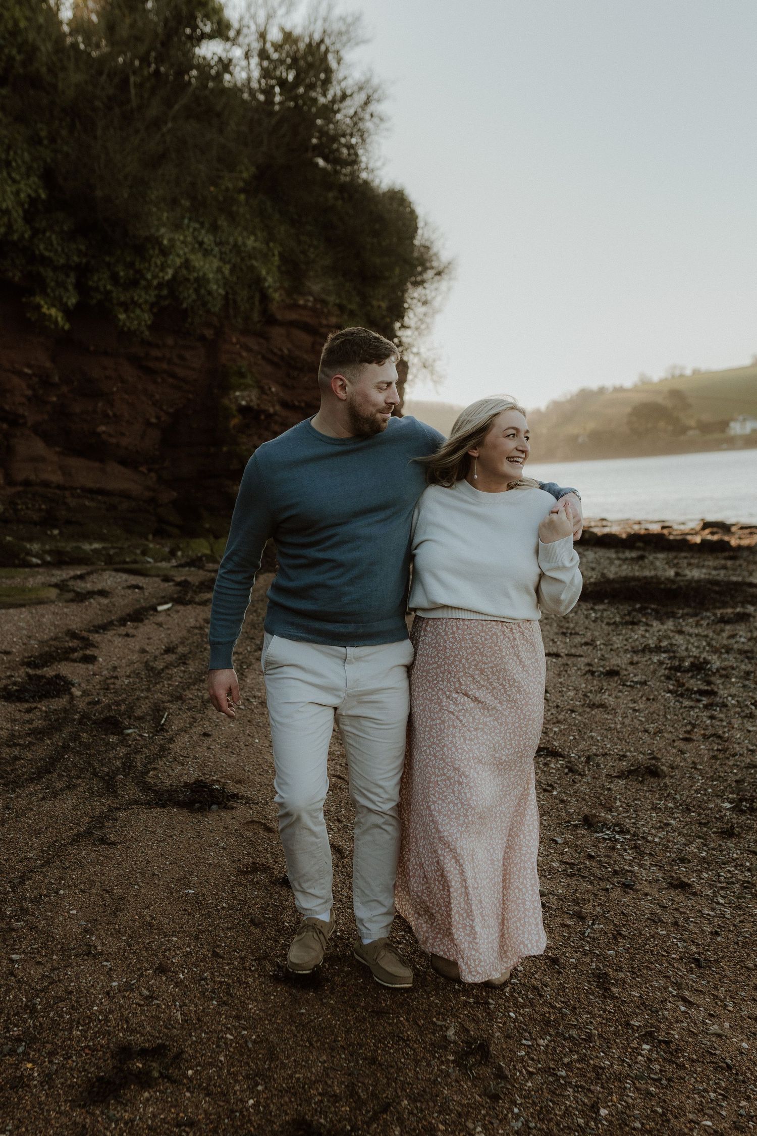 A couple walks together along a rocky beach shoreline at sunset in matching casual outfits.