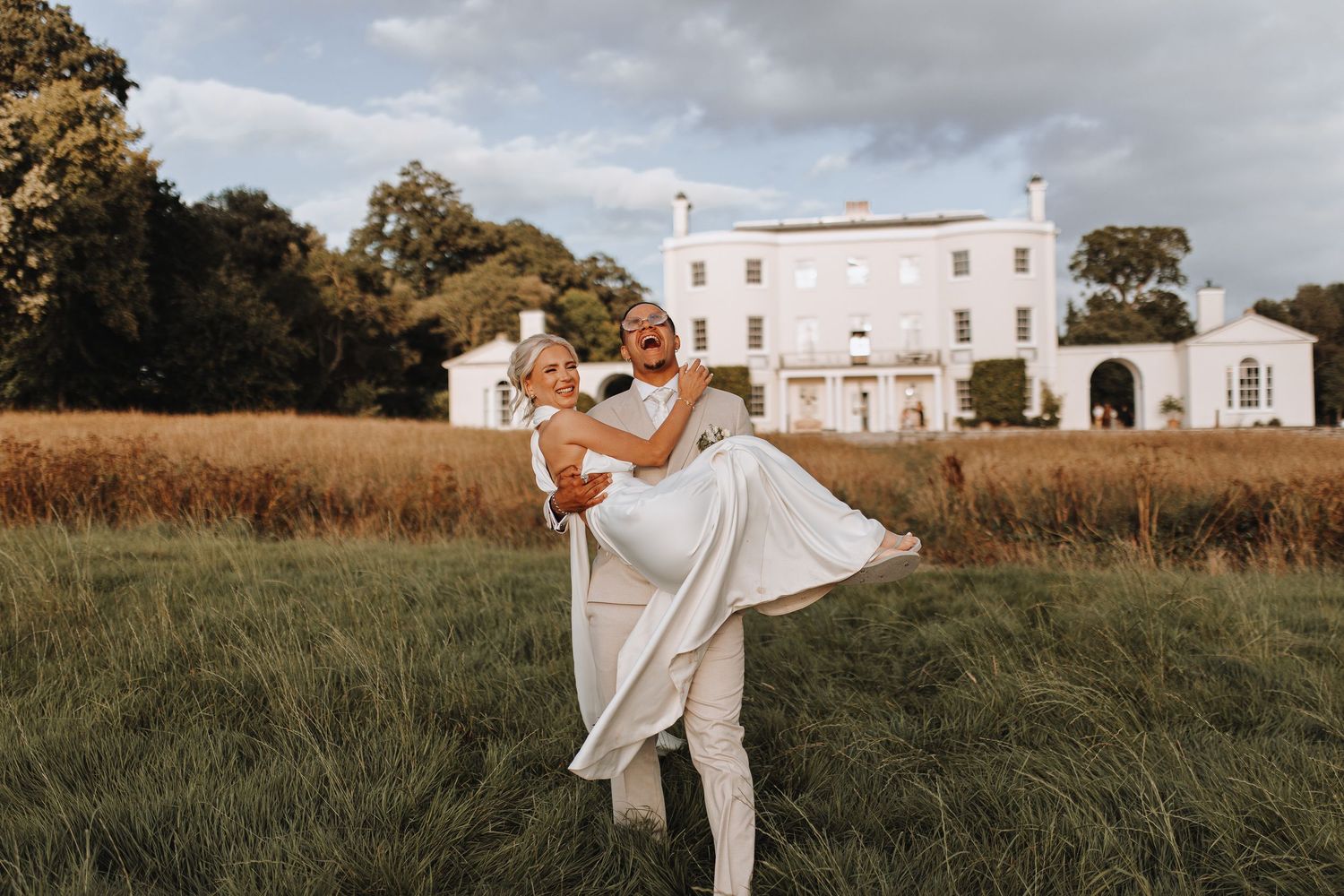 A romantic couple dances together in a grassy field at sunset with a white mansion in the background.