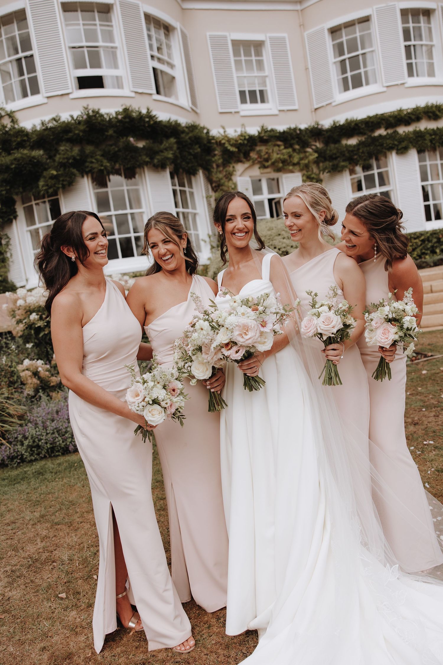 A wedding party in blush pink dresses stands together laughing and holding white floral bouquets on a grassy lawn.