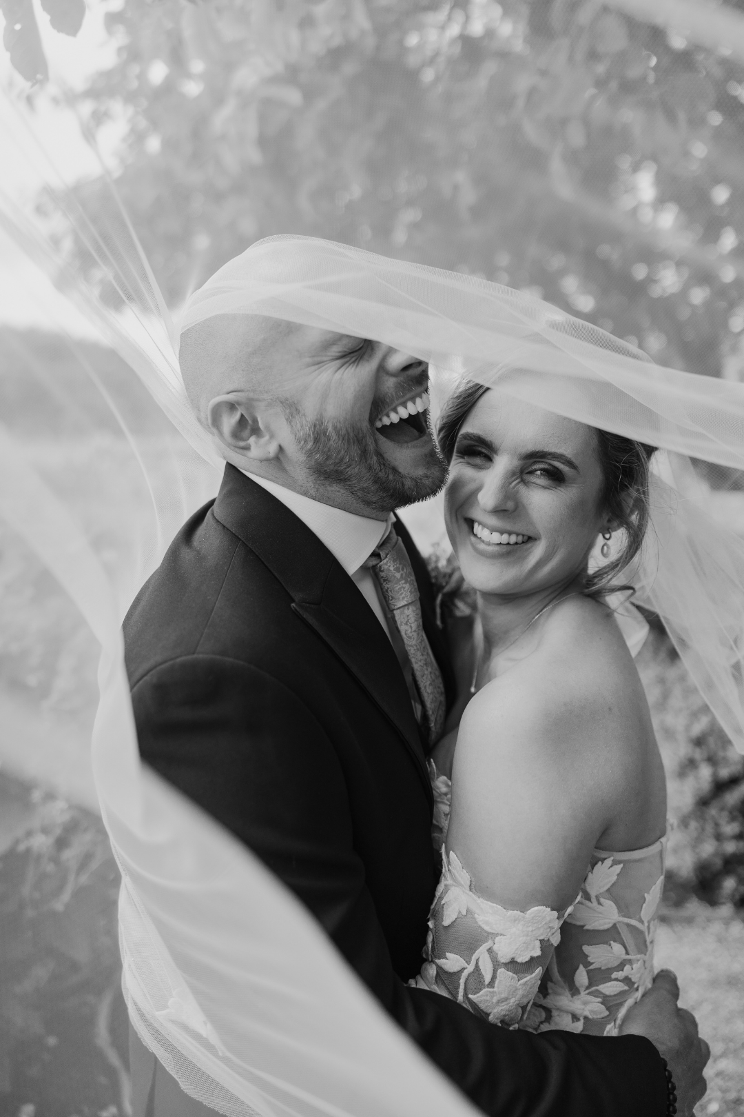 A joyful bridal couple shares a tender moment under a flowing veil in black and white photography.