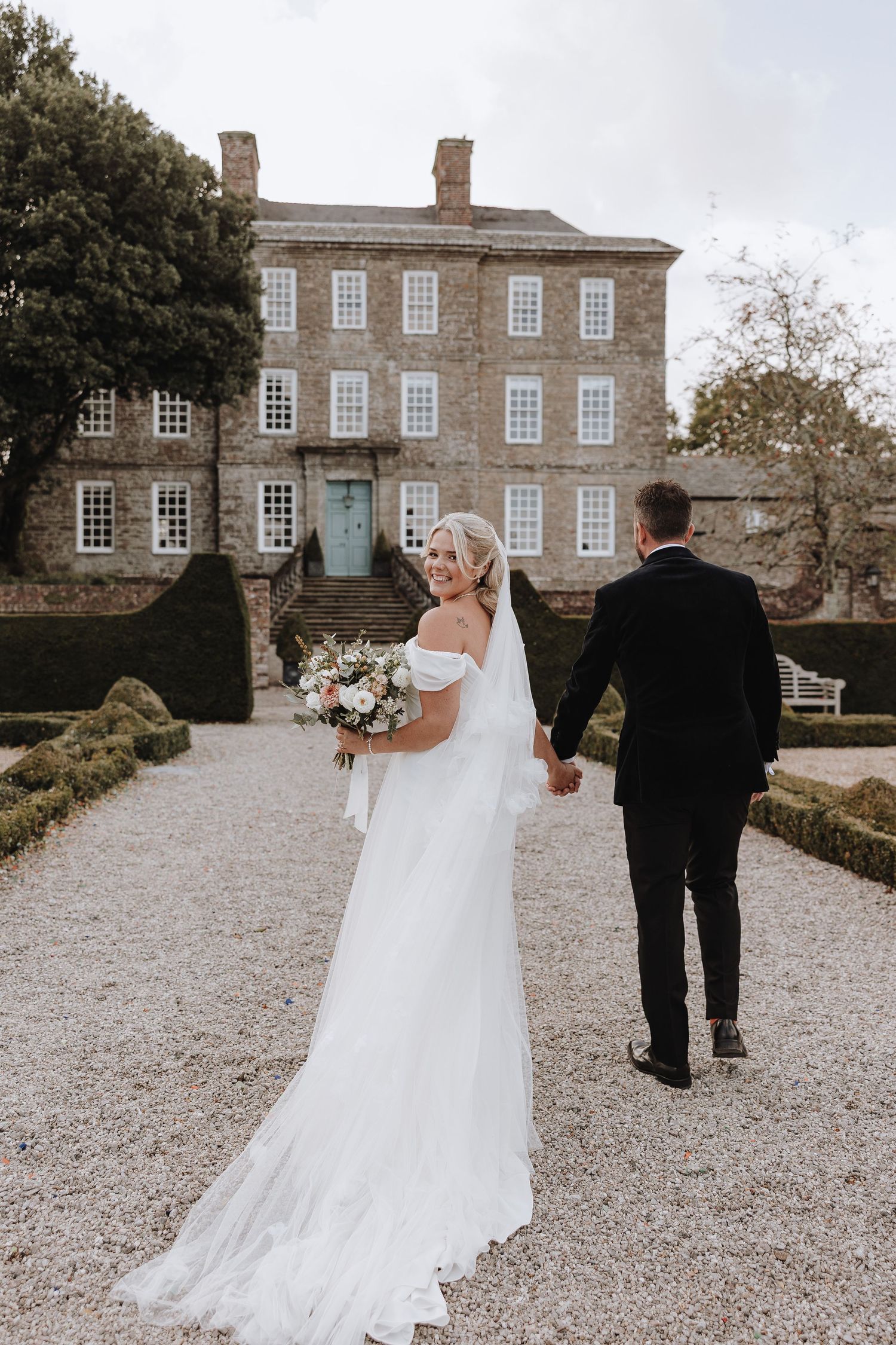 A couple walks along a gravel path towards a stately Georgian manor house on their wedding day.