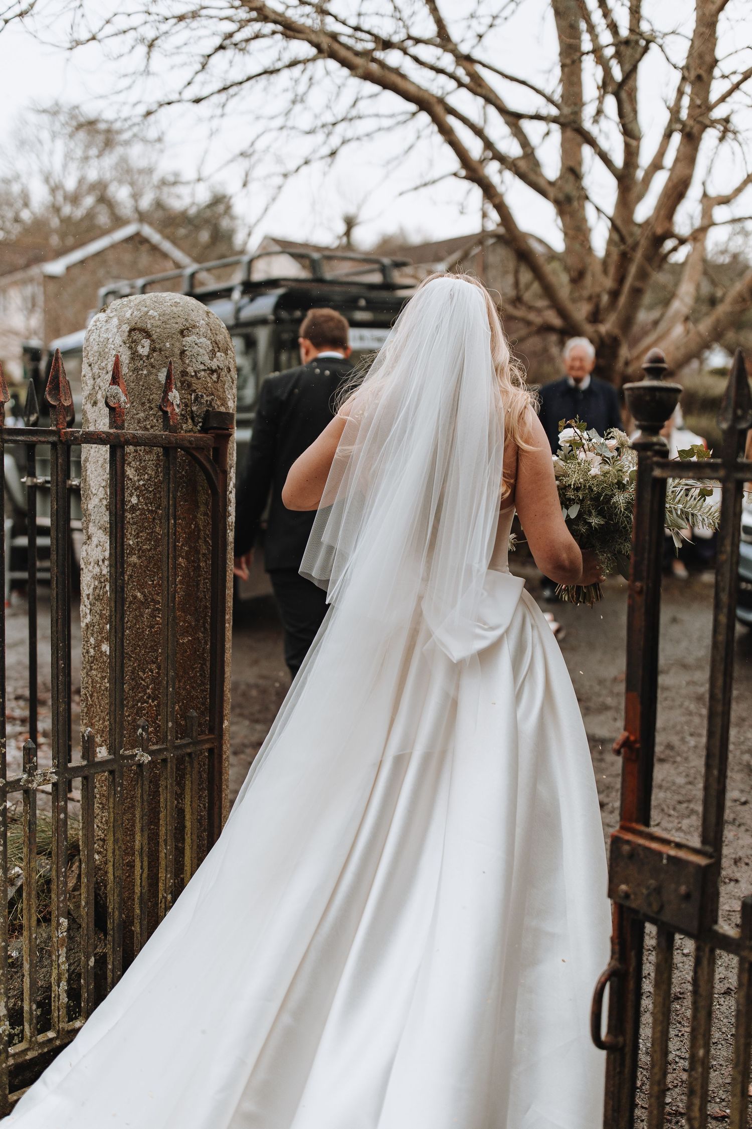 A flowing white wedding dress train trails behind as it moves through a rustic iron gate in an outdoor setting.