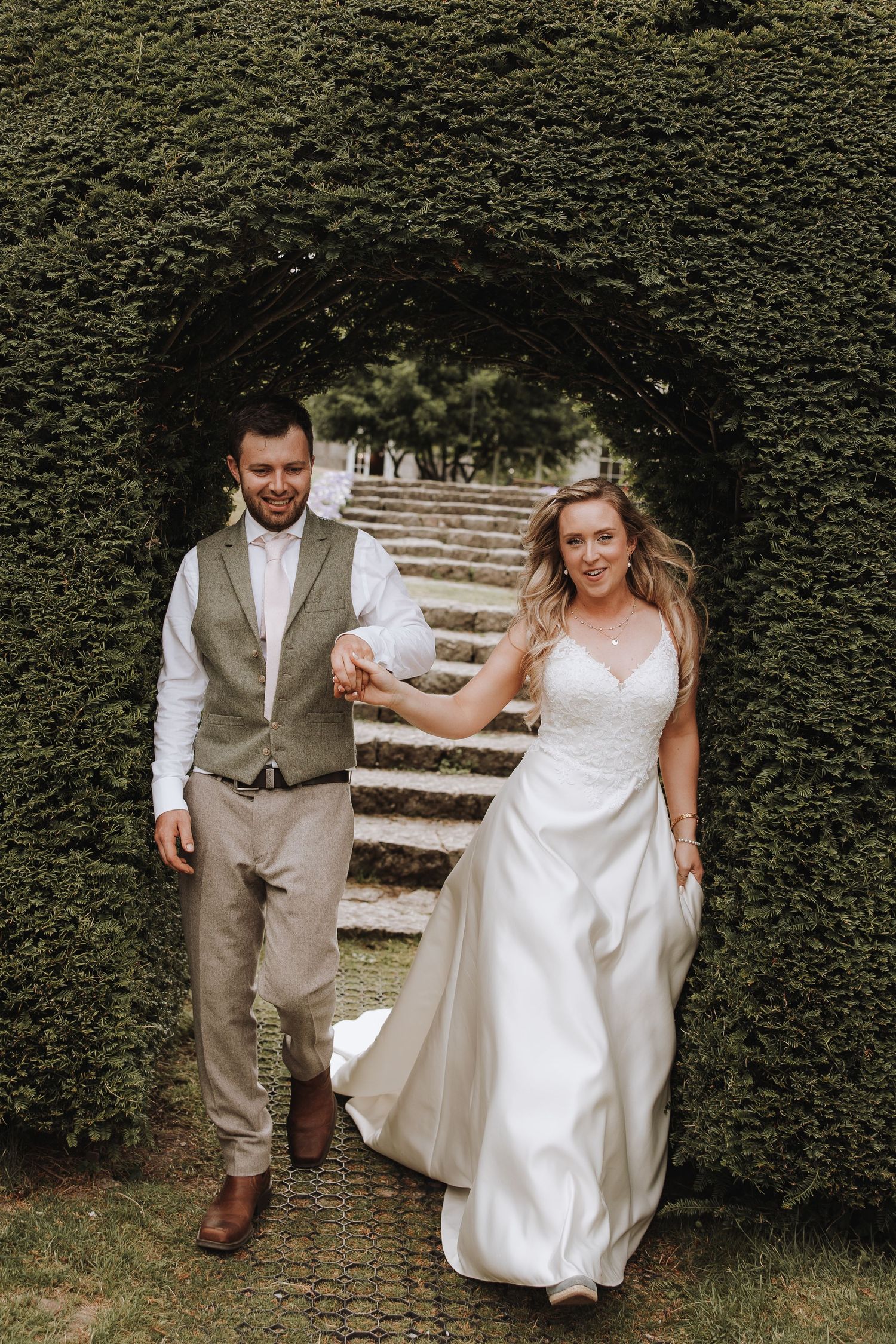 A couple in wedding attire walking together on stone steps surrounded by lush green hedges.