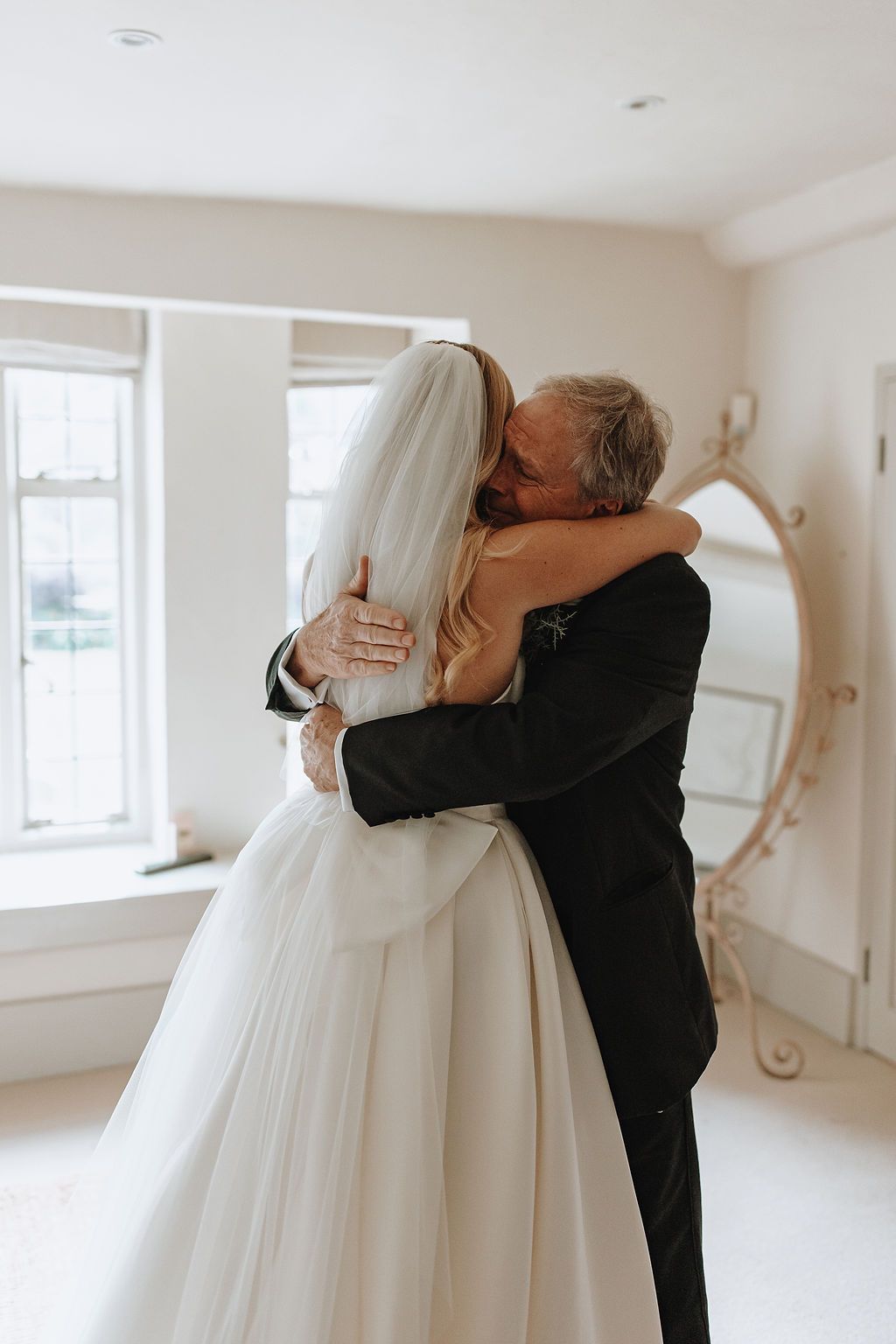 An emotional embrace between two people at a wedding ceremony in a bright white room with large windows.