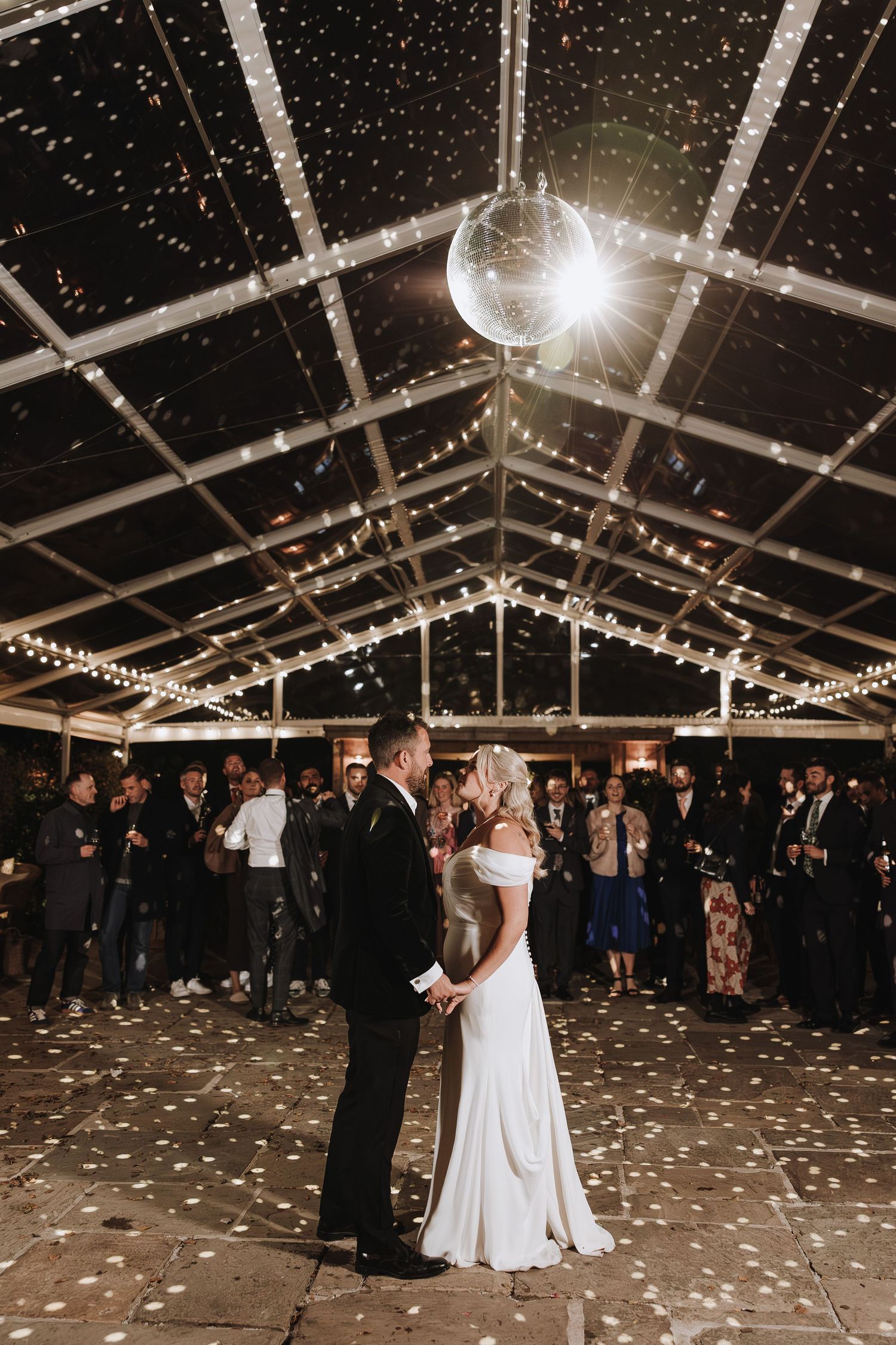 A couple shares their first dance under a clear tent decorated with twinkling lights and scattered petals at their wedding reception.