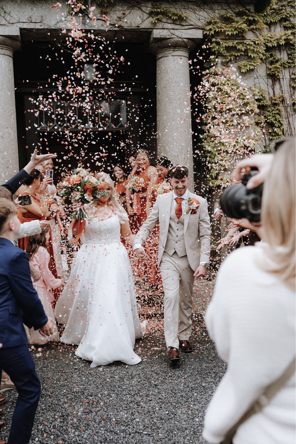 Bride and groom exit ceremony venue in confetti shower, surrounded by guests in orange attire, as photographer captures the moment.