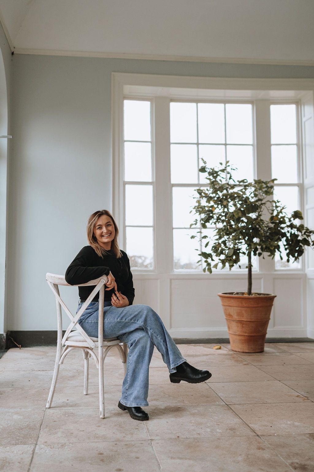 A person sits casually on a white bistro chair by large windows with a potted tree in a bright, minimalist interior.