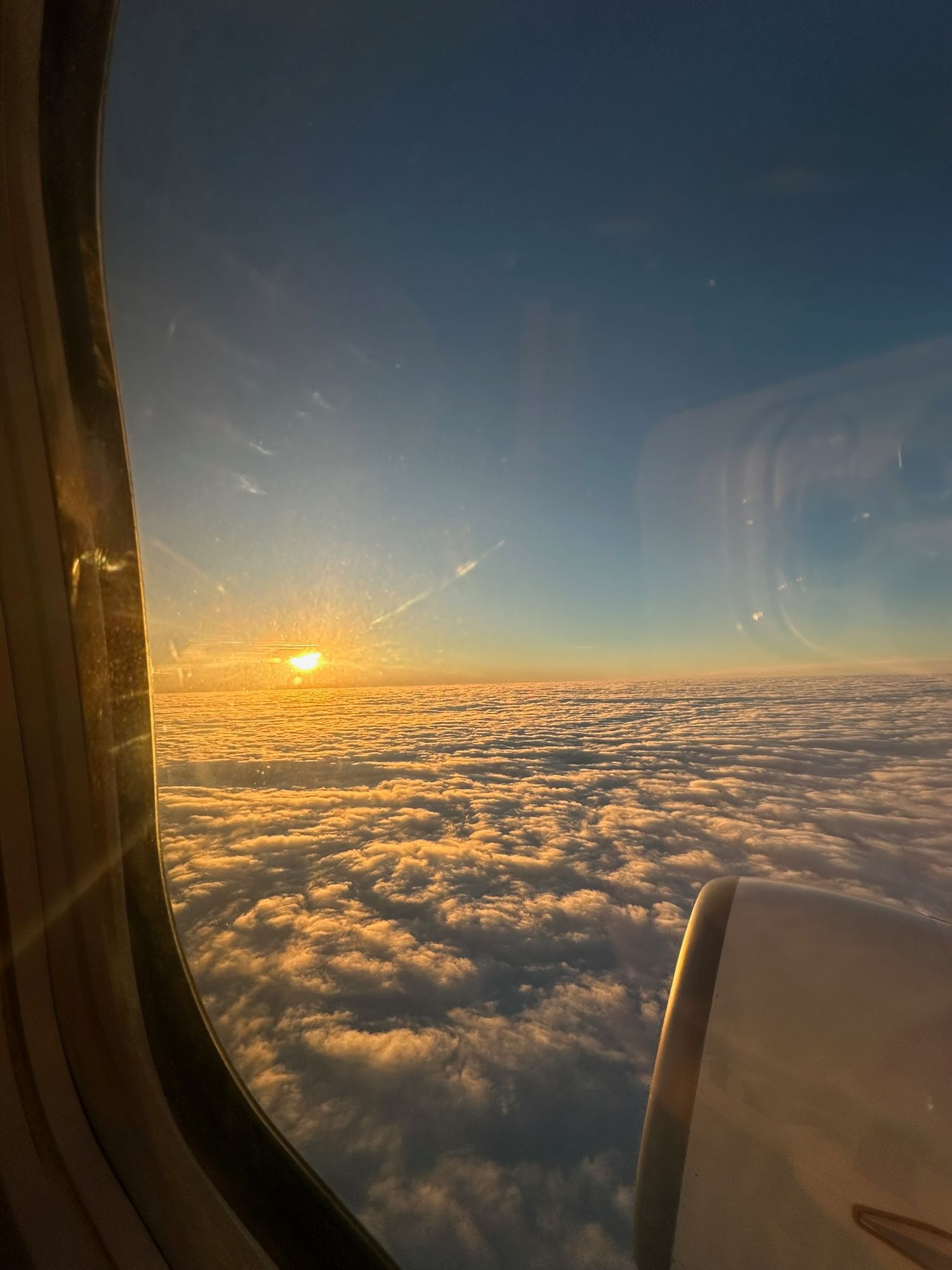 Airplane window view of sunrise over fluffy clouds at high altitude with golden light illuminating the horizon.