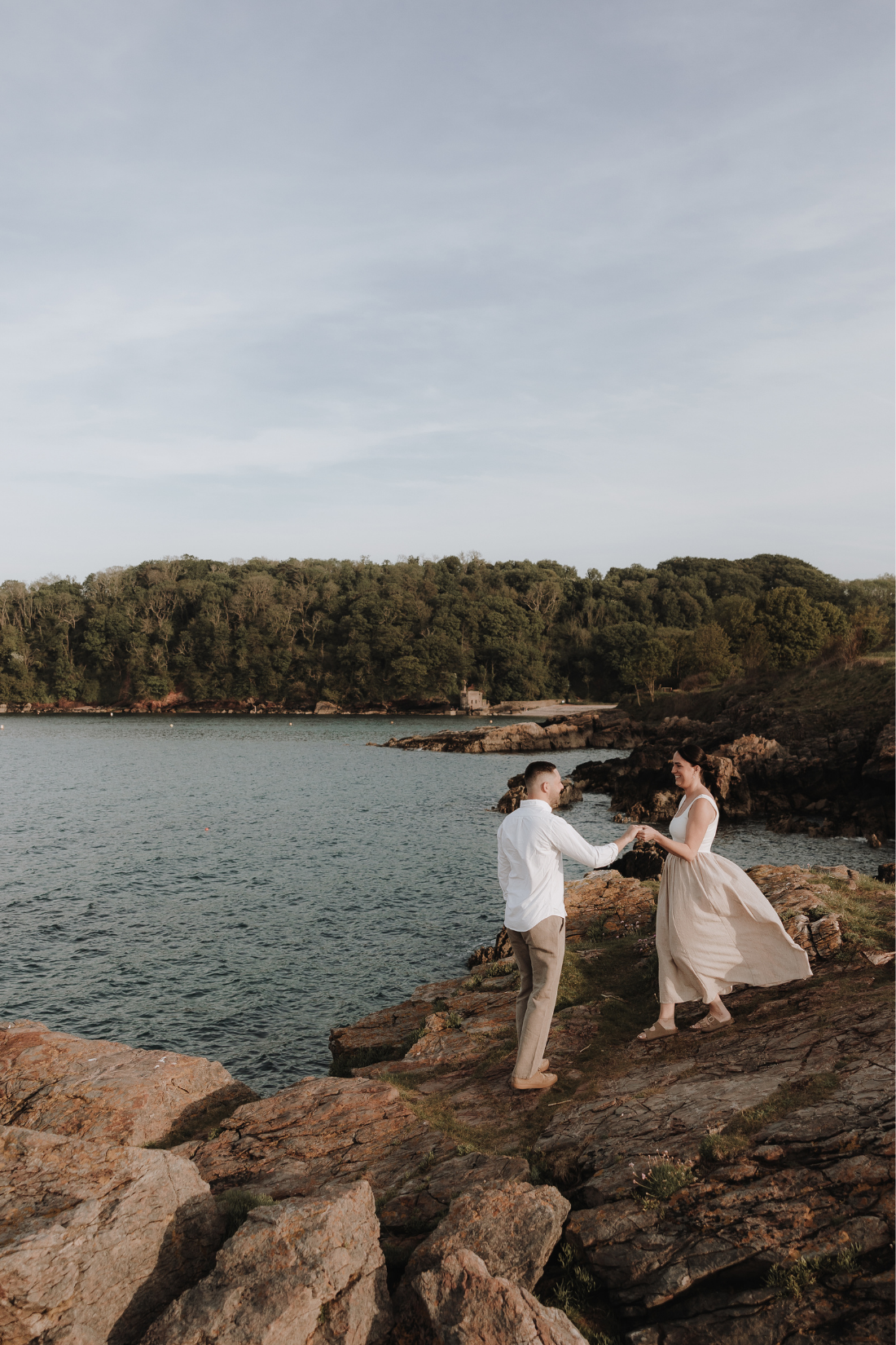 Couple holding hands on rocky coastal cliffs with turquoise water and lush green trees in the background.