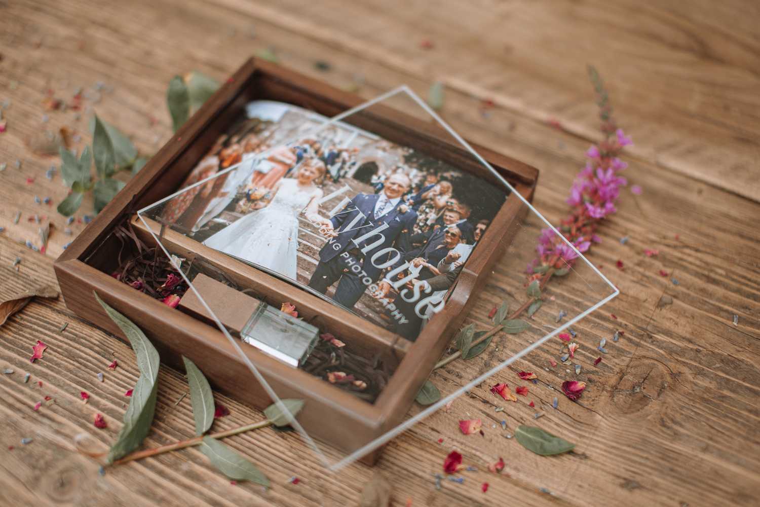 A wedding photo frame with a group shot sits on a rustic wooden surface decorated with eucalyptus leaves and pink flowers.