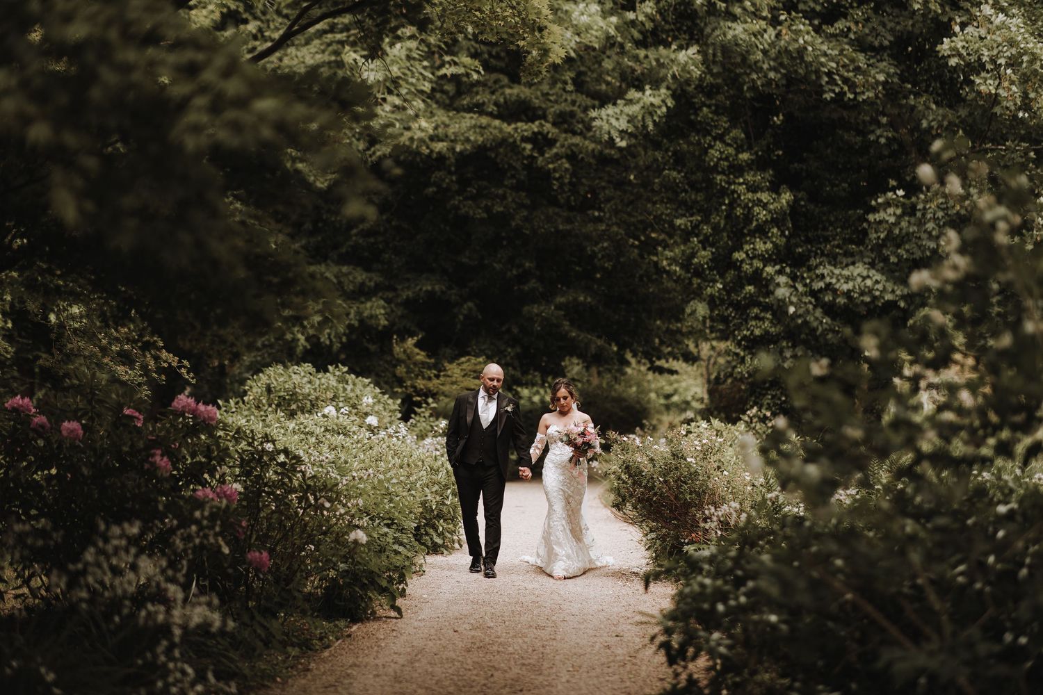 A couple walks down a winding garden path at Deer Park Country House in Honiton surrounded by lush greenery and blooming flowers.