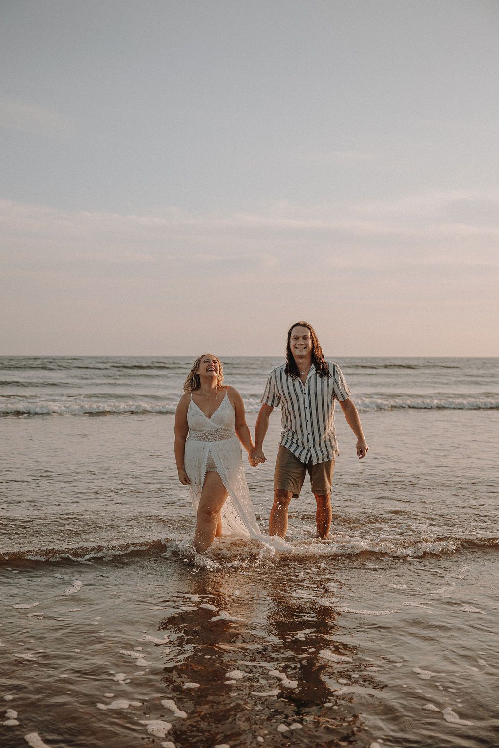A couple splashes together in shallow ocean waves at sunset while wearing casual beach attire.