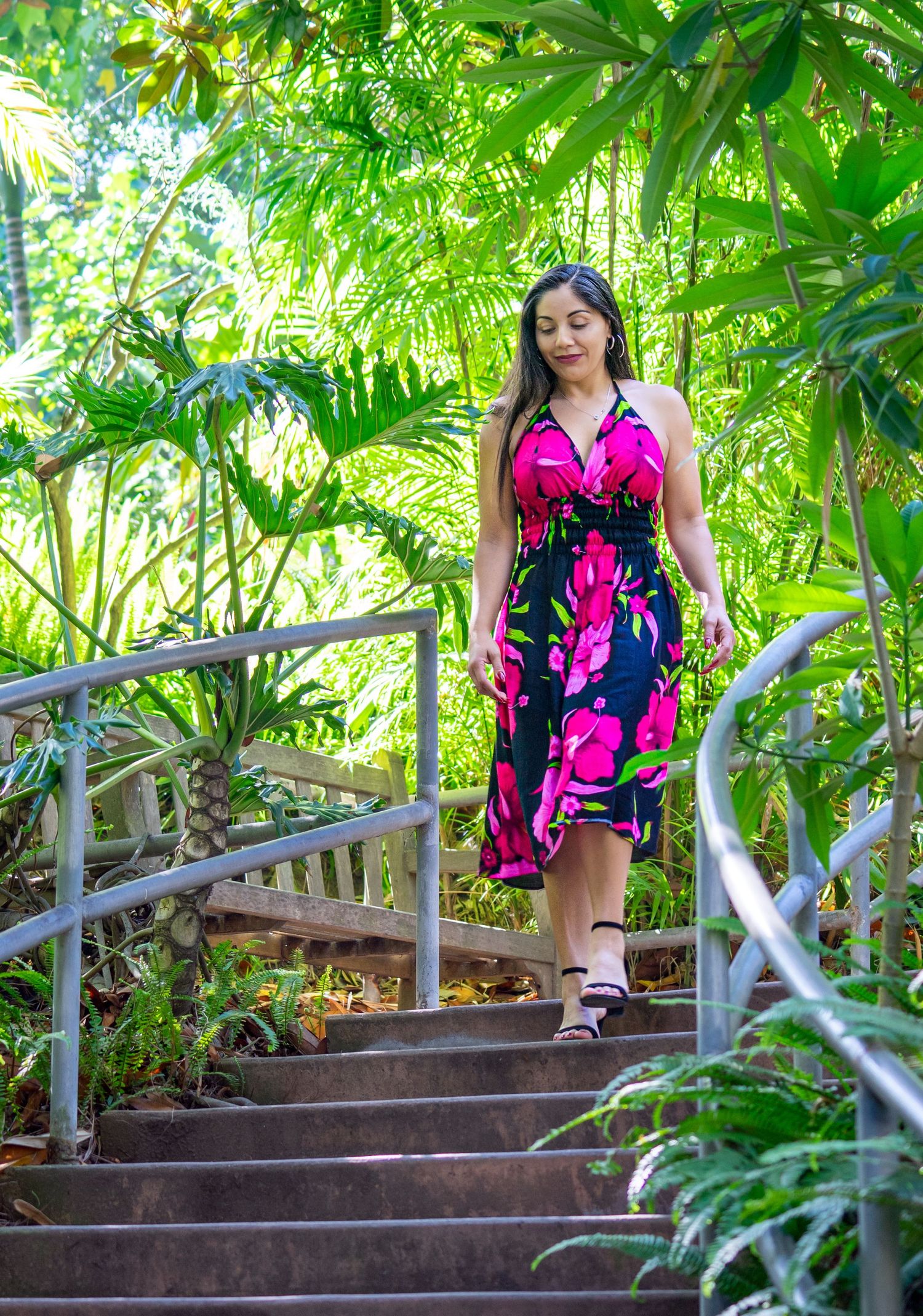 Woman in bright pink floral dress descends stone steps surrounded by lush tropical foliage.
