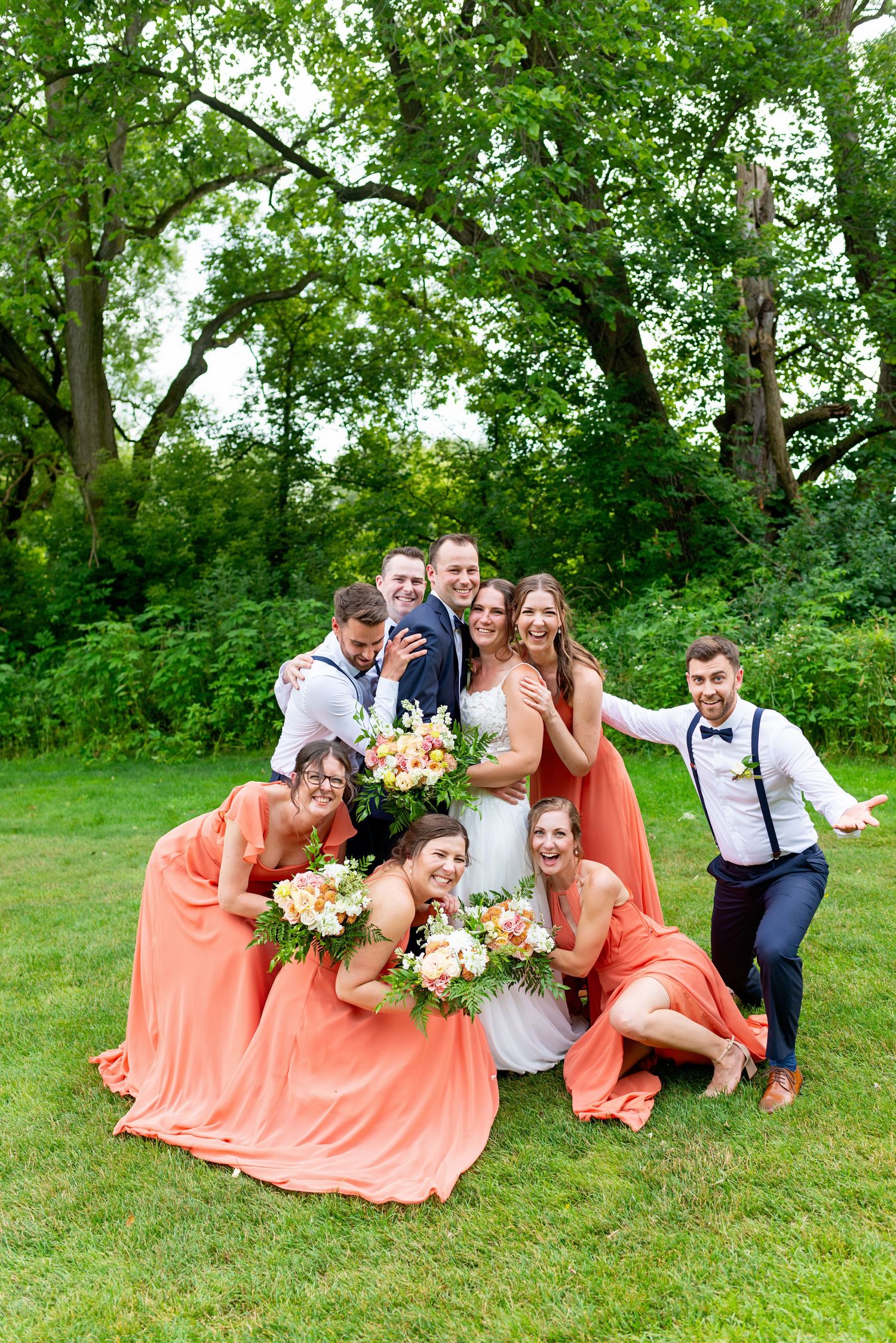 Wedding party in coral bridesmaid dresses and navy suits posing playfully outdoors on green grass with lush trees.