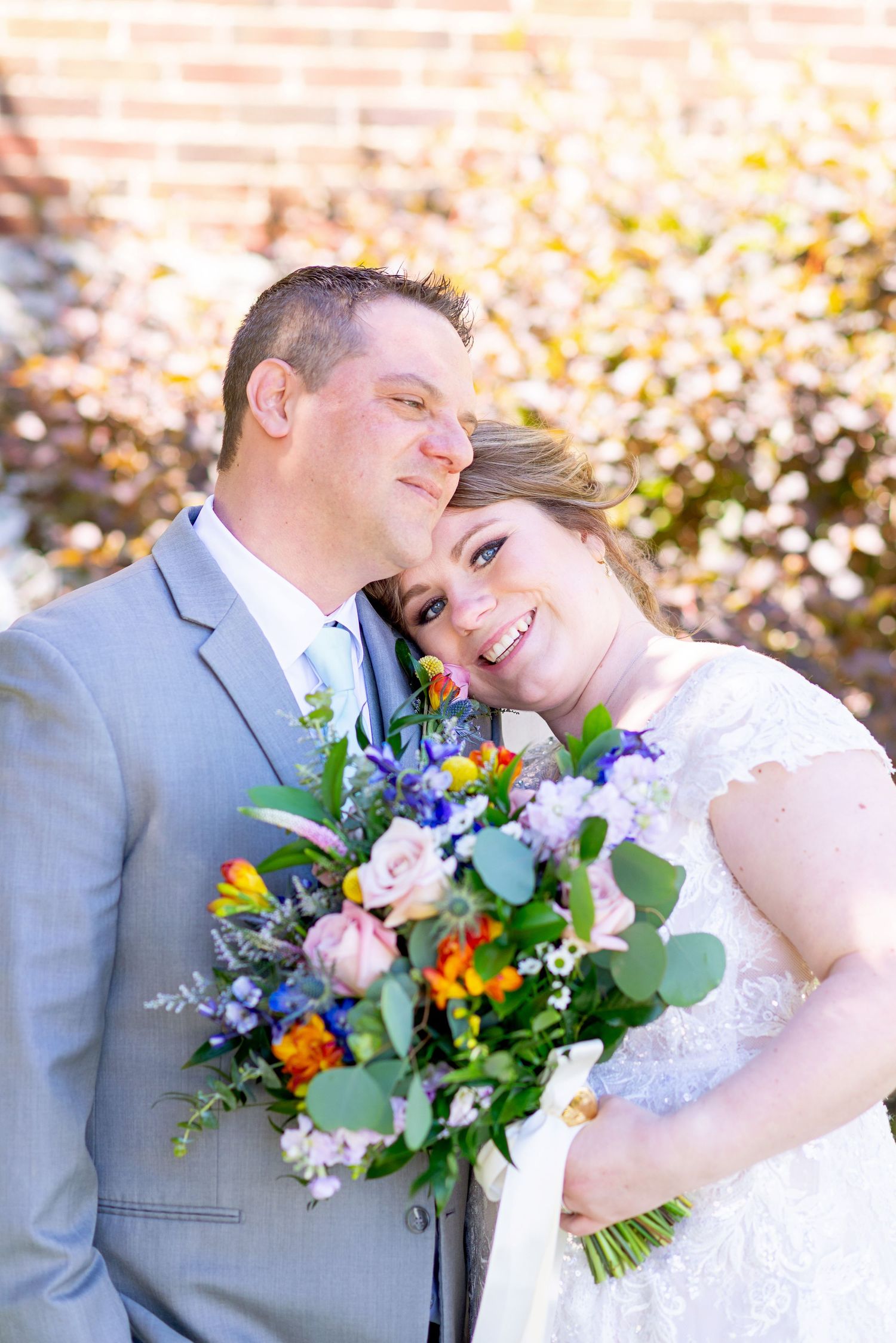 Smiling bride holding colorful bouquet leans against groom in gray suit outdoors at wedding.
