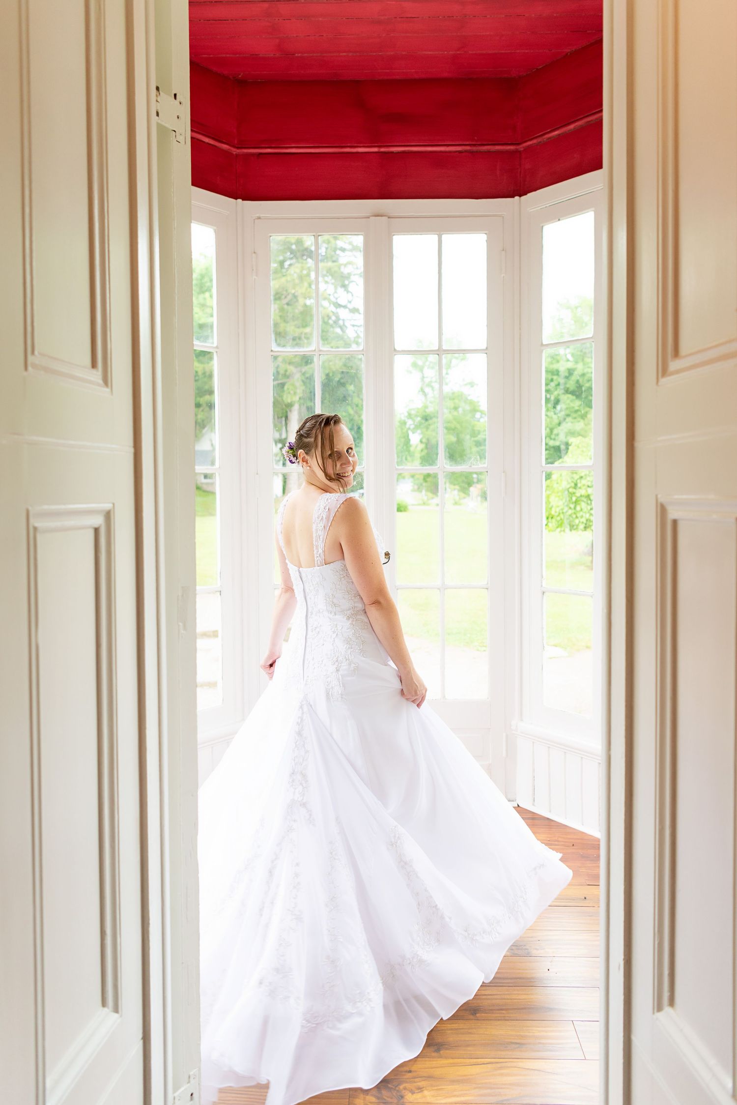 Bride in white wedding gown standing in elegant doorway with red ceiling, facing large windows overlooking green gardens.