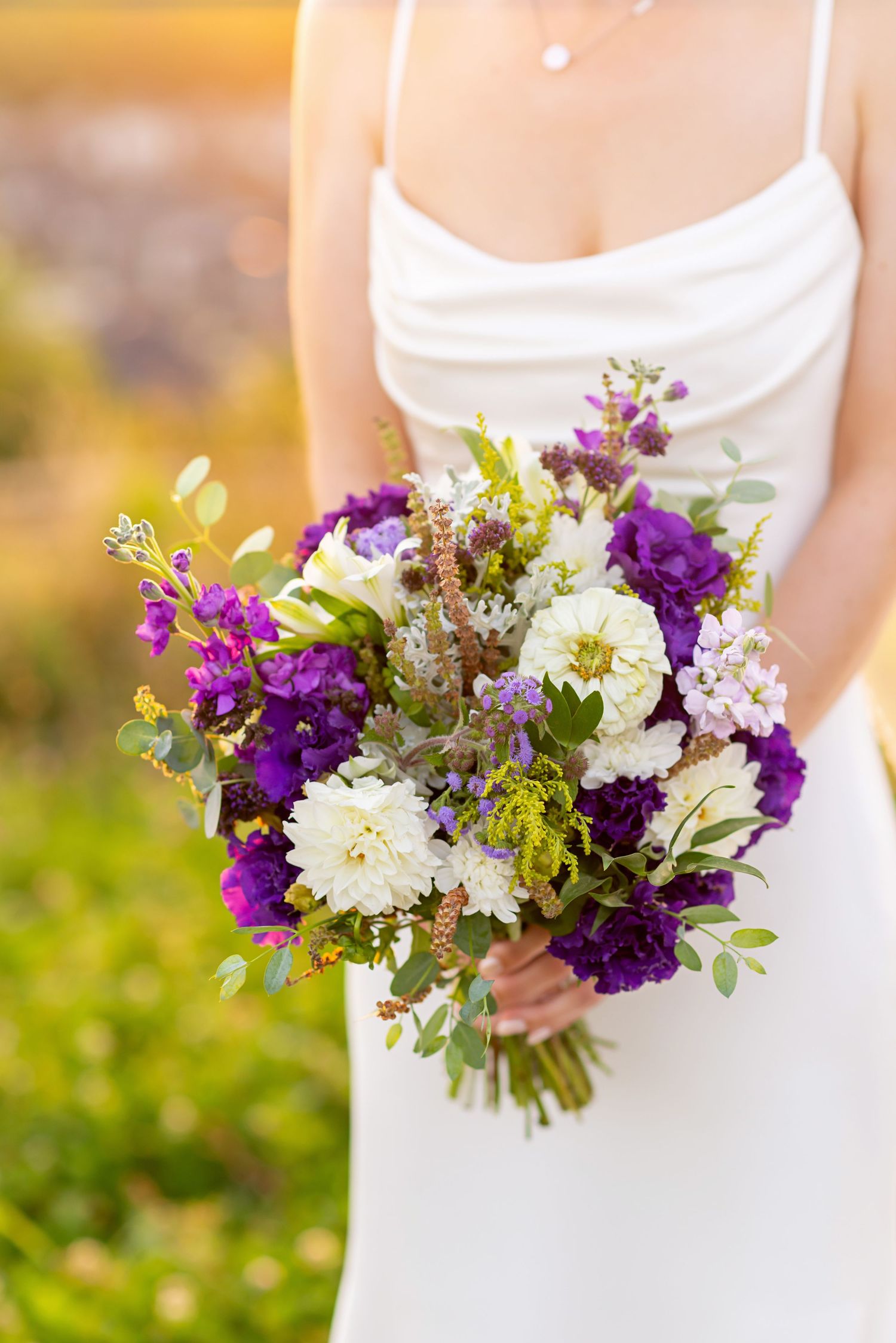 Bride in white gown holding a lush purple and white wedding bouquet with eucalyptus greenery outdoors.