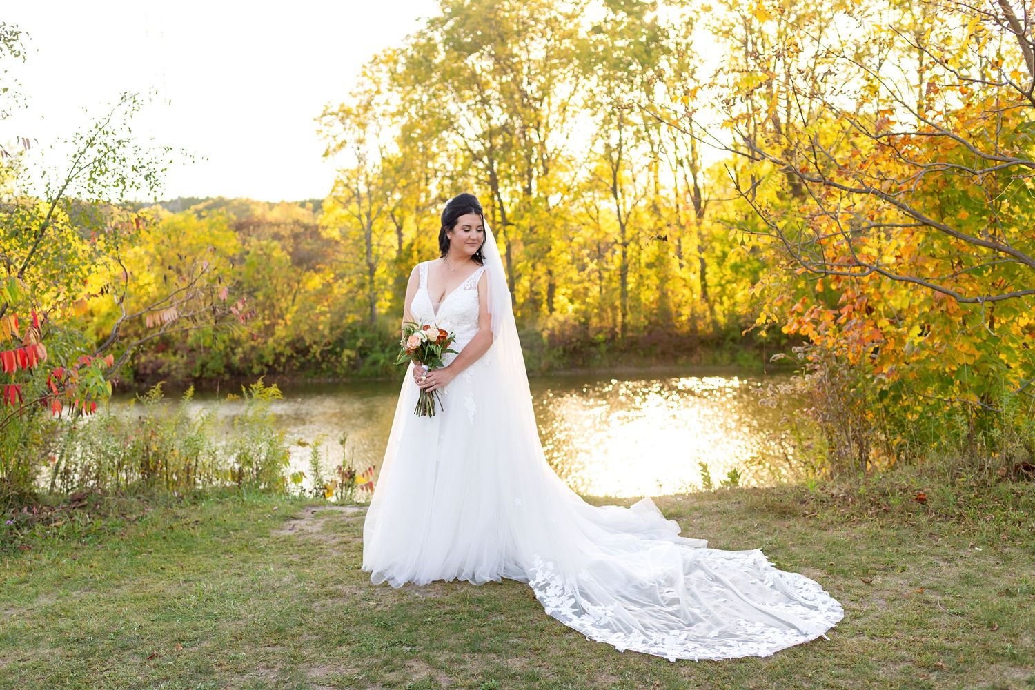 Bride in white gown with long train holds bouquet by autumn lake with colorful fall foliage backdrop.