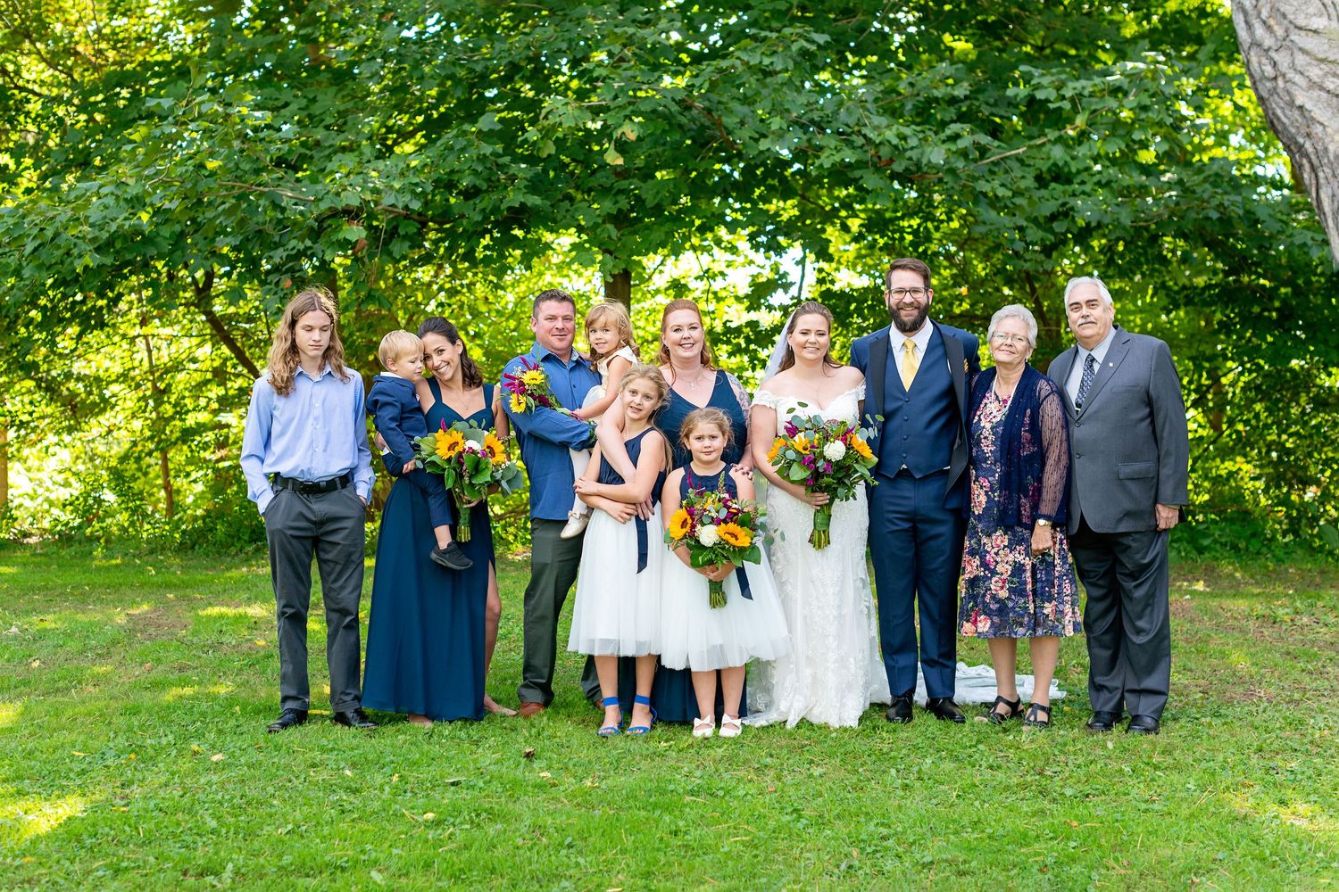 Wedding party poses outdoors on green lawn, bride in white gown, bridesmaids in navy blue dresses, holding colorful bouquets.