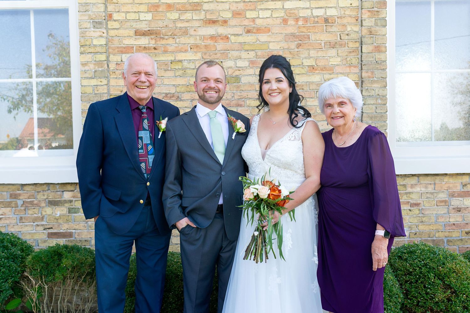 Bride and groom pose with elderly couple outside brick building on wedding day, bride holding colorful bouquet.