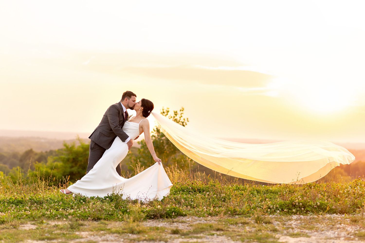 Bride and groom share a romantic kiss at sunset, with her veil flowing in the wind on a scenic hilltop.