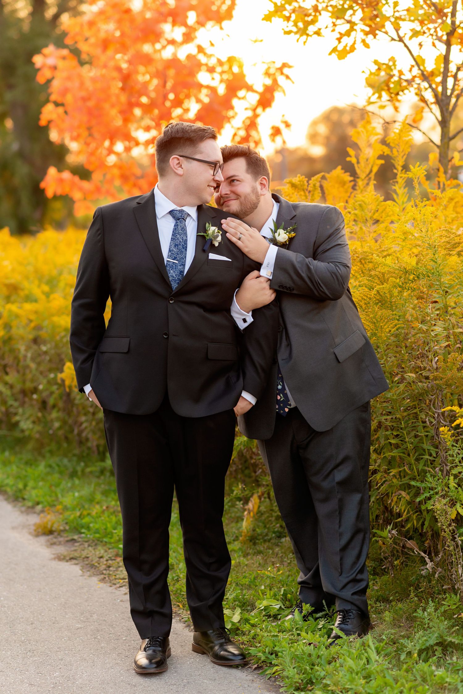 Two grooms in dark suits share a romantic moment on a path surrounded by vibrant autumn foliage.