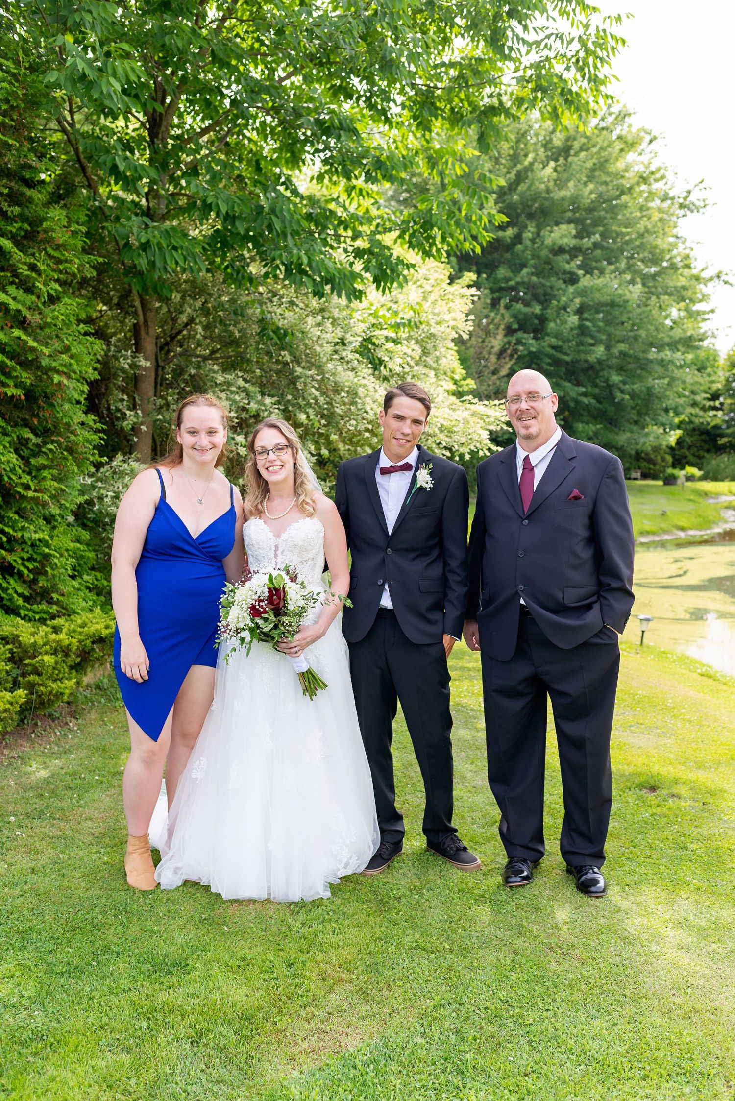 Bride and groom pose with family on green lawn, bride holding bouquet, guests in formal attire with trees in background.