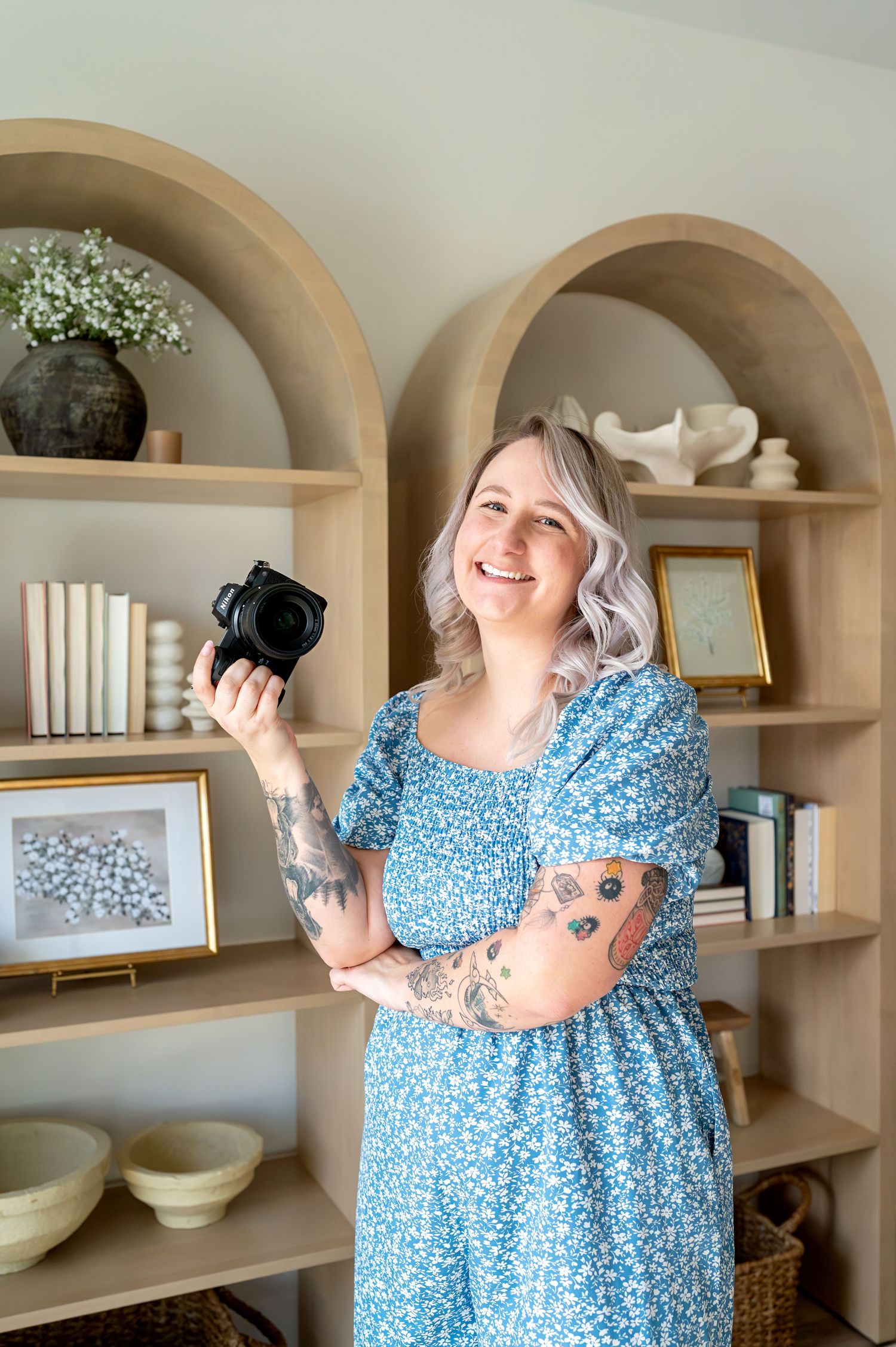 Smiling woman with lavender hair holding a camera in a blue floral dress, standing in front of wooden arch shelving.