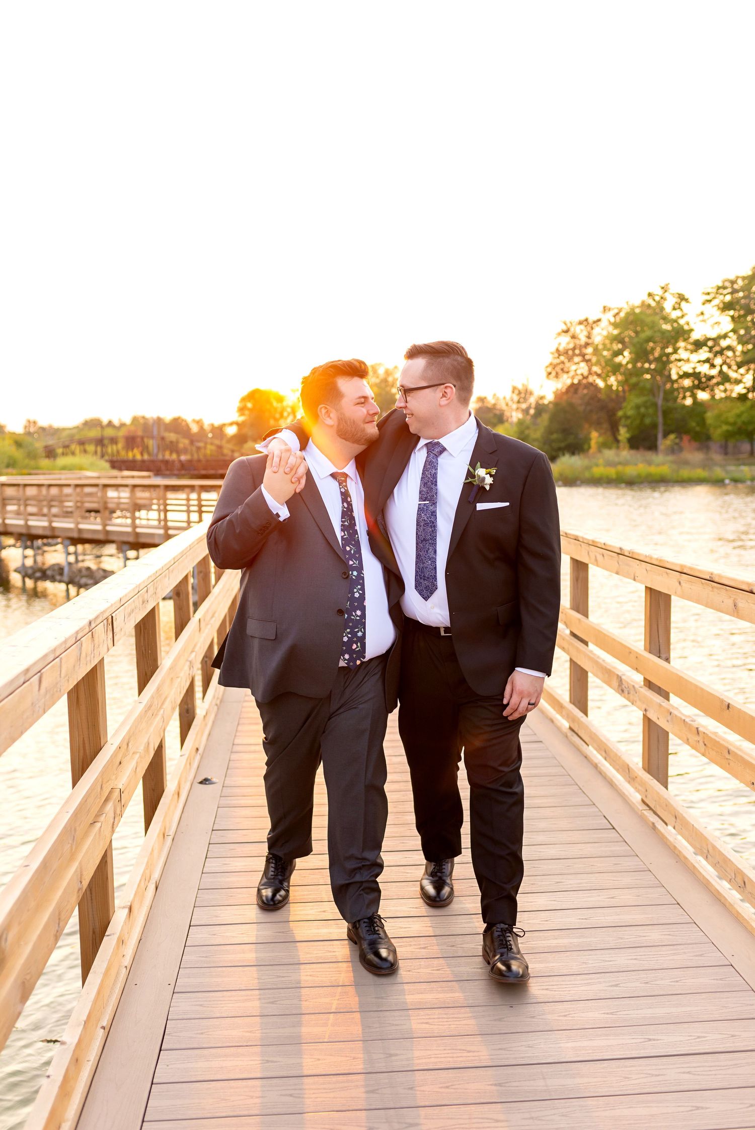 Two grooms in suits walk hand in hand on a wooden dock at sunset, sharing a romantic moment on their wedding day.