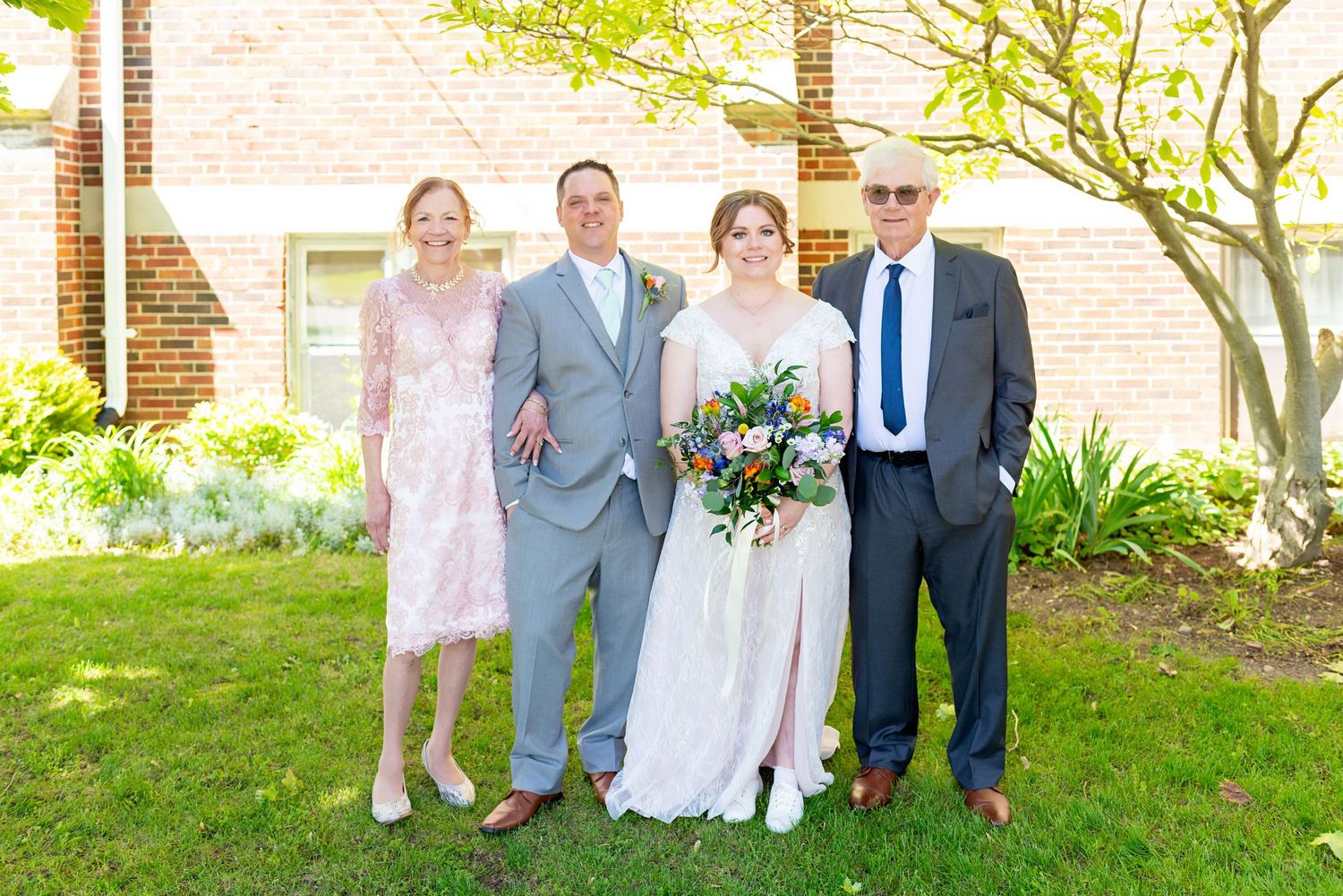 Wedding party of four posing outdoors, bride holding colorful bouquet, groom in gray suit, beside brick building.