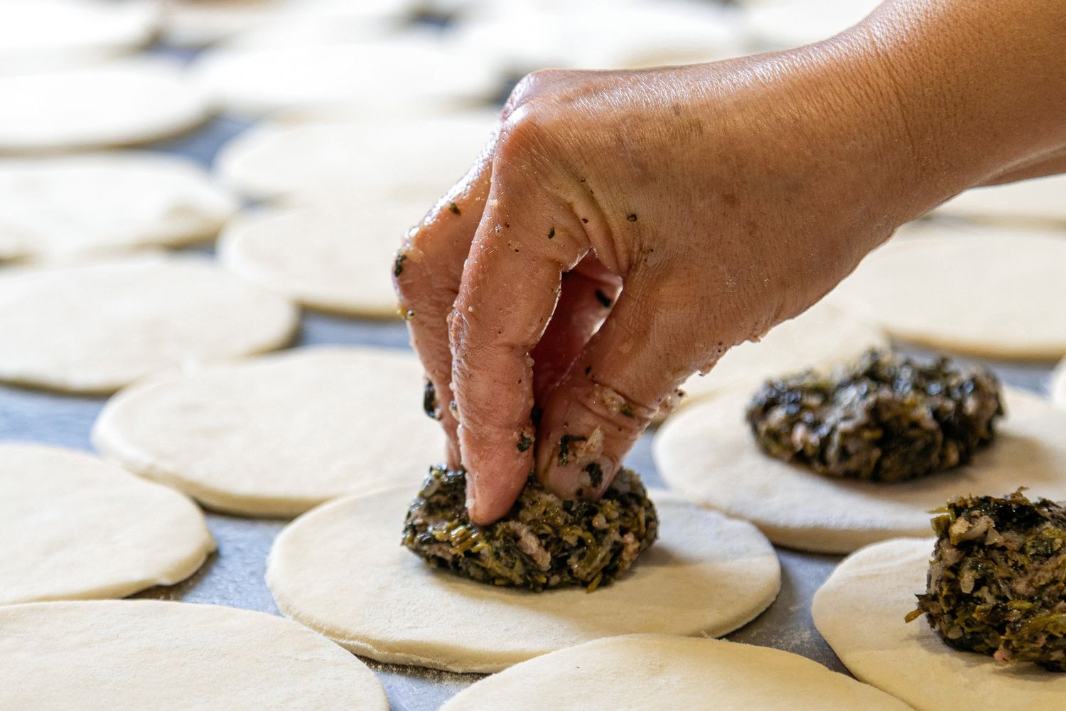 Hands placing spinach filling onto round dumpling wrappers laid out in rows on a white surface.