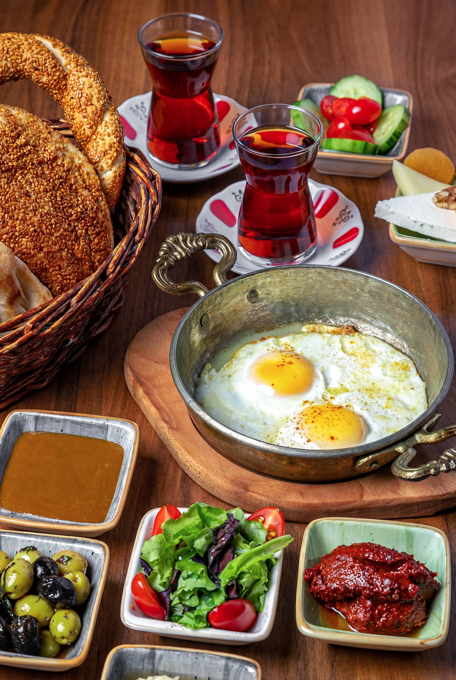 Traditional Turkish breakfast spread with fried eggs, simit bread, tea in tulip glasses, olives and fresh vegetables on wooden table.
